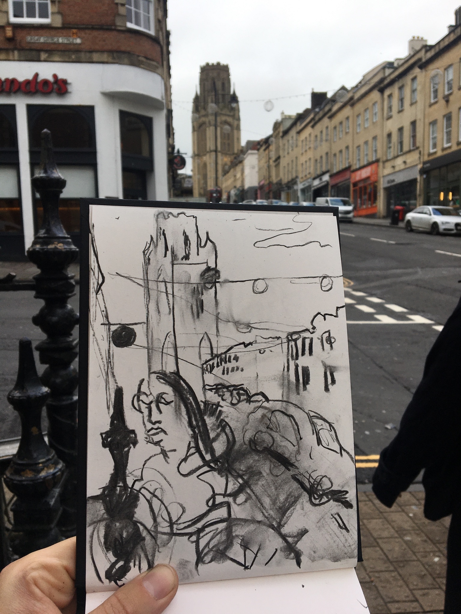 A photograph of a hand holding an open sketchbook in front of a city street. The sketchbook has a charcoal drawing of the street on the open page.