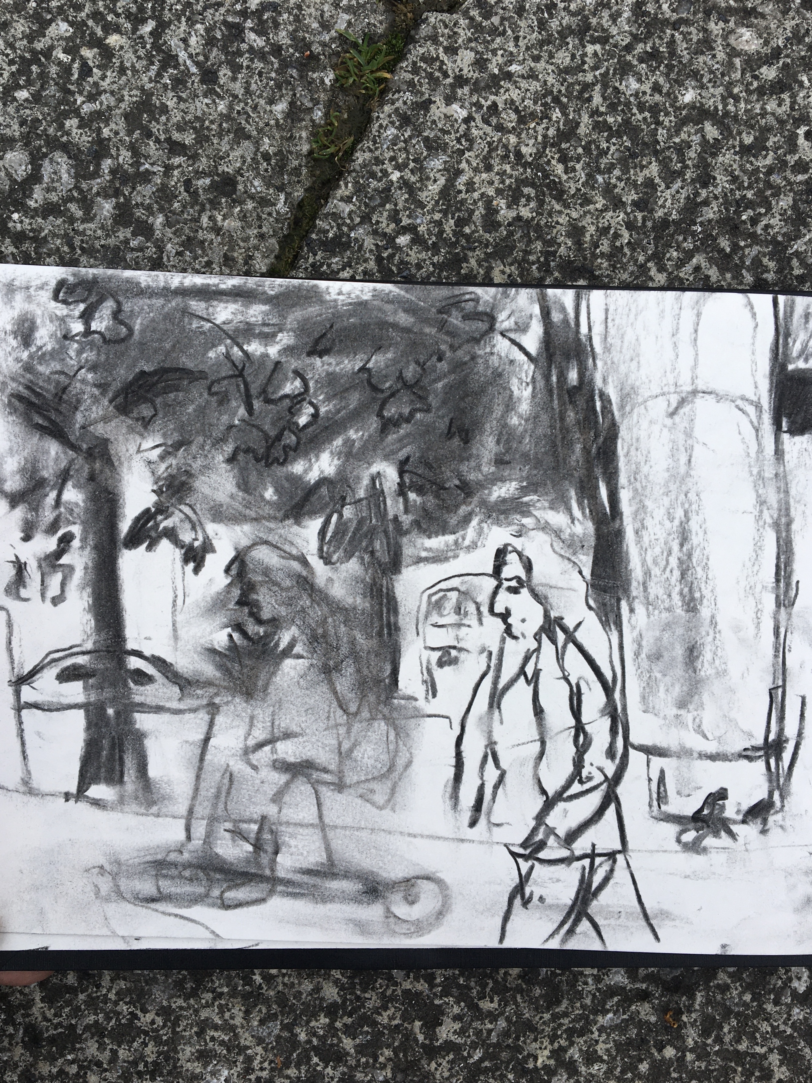 A photograph of an open sketchbook placed on a pavement. The sketchbook has a charcoal drawing of a street scene on the open page.
