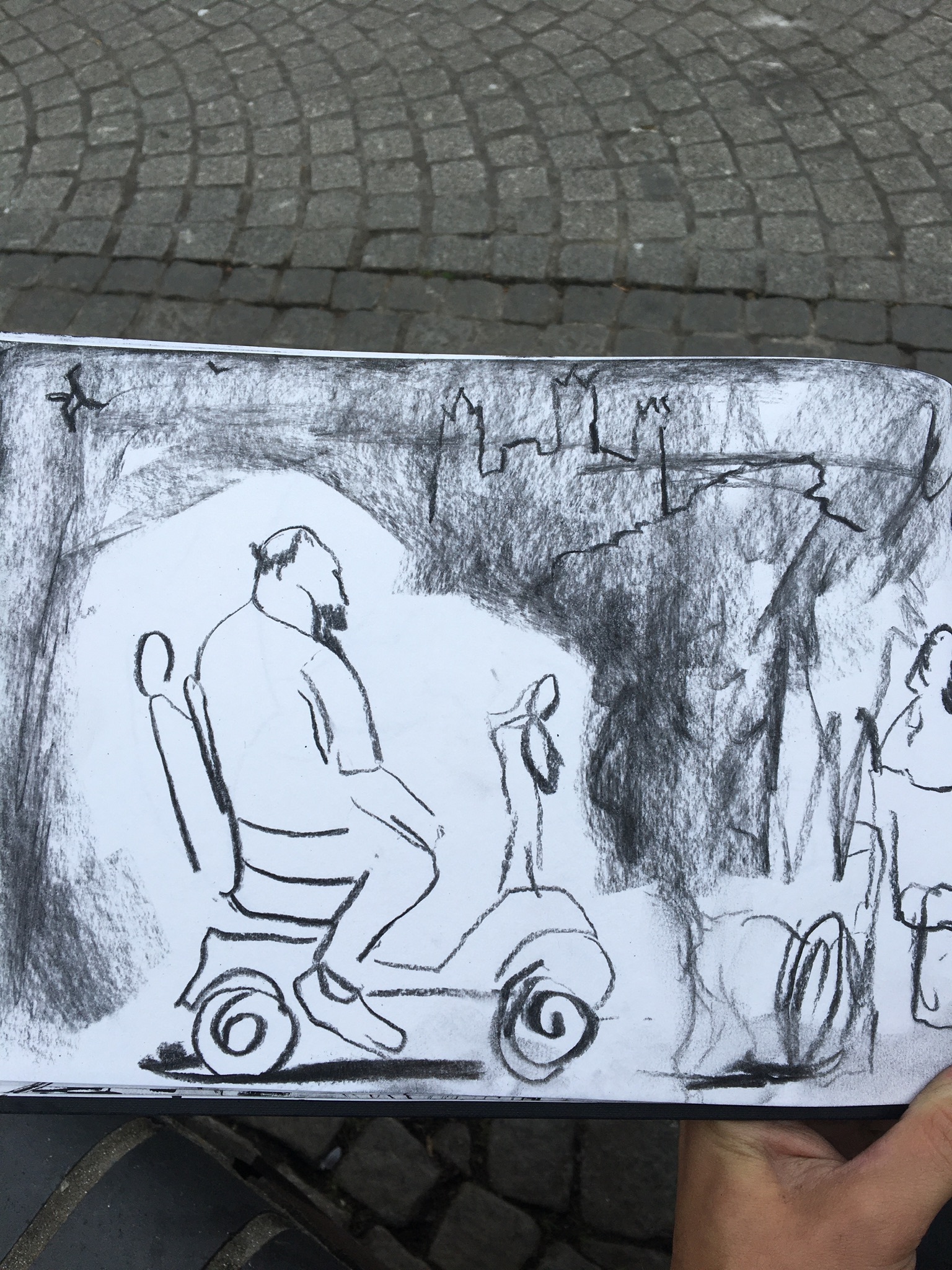 A photograph of an open sketchbook placed on a pavement. The sketchbook has a charcoal drawing of a street scene on the open page, featuring a person an a mobility scooter..