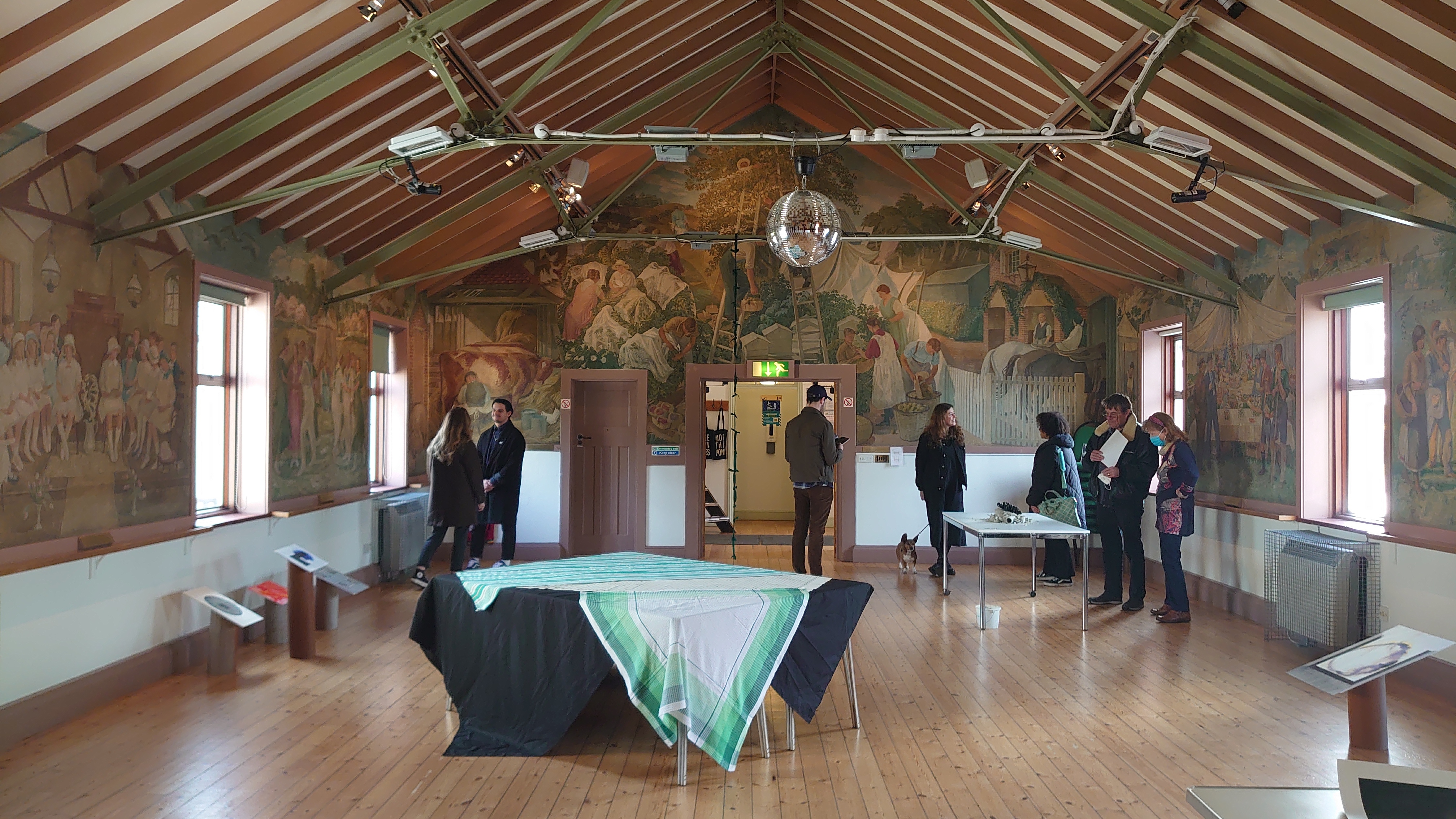 A small group of people are standing in a room with wooden beams and decorative wallpapered walls. There is a table in the centre if the images with a colourful tablecloth draped over it and a disco ball hanging from the ceiling