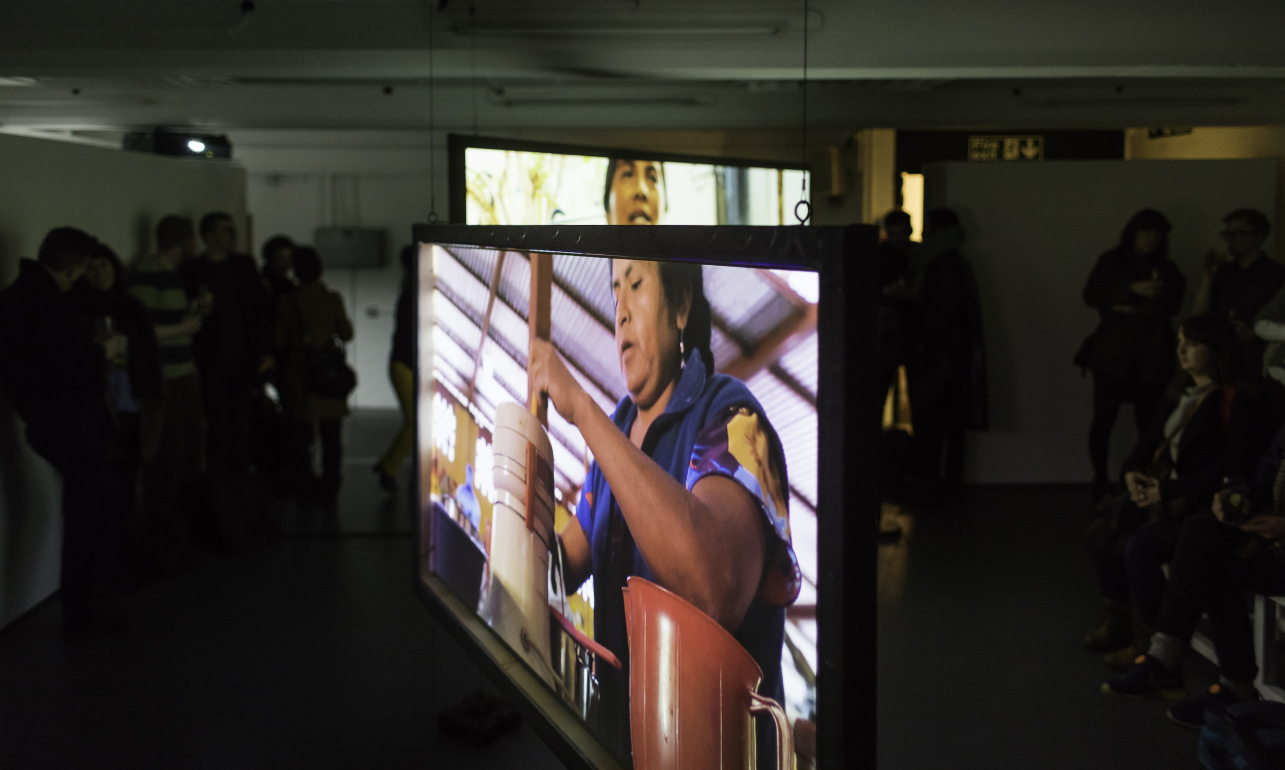 A large hanging screen shows a projection of a woman mixing something. In the background is the top edge of another screen and in the darkened gallery space we can see crowds of people watching the two films