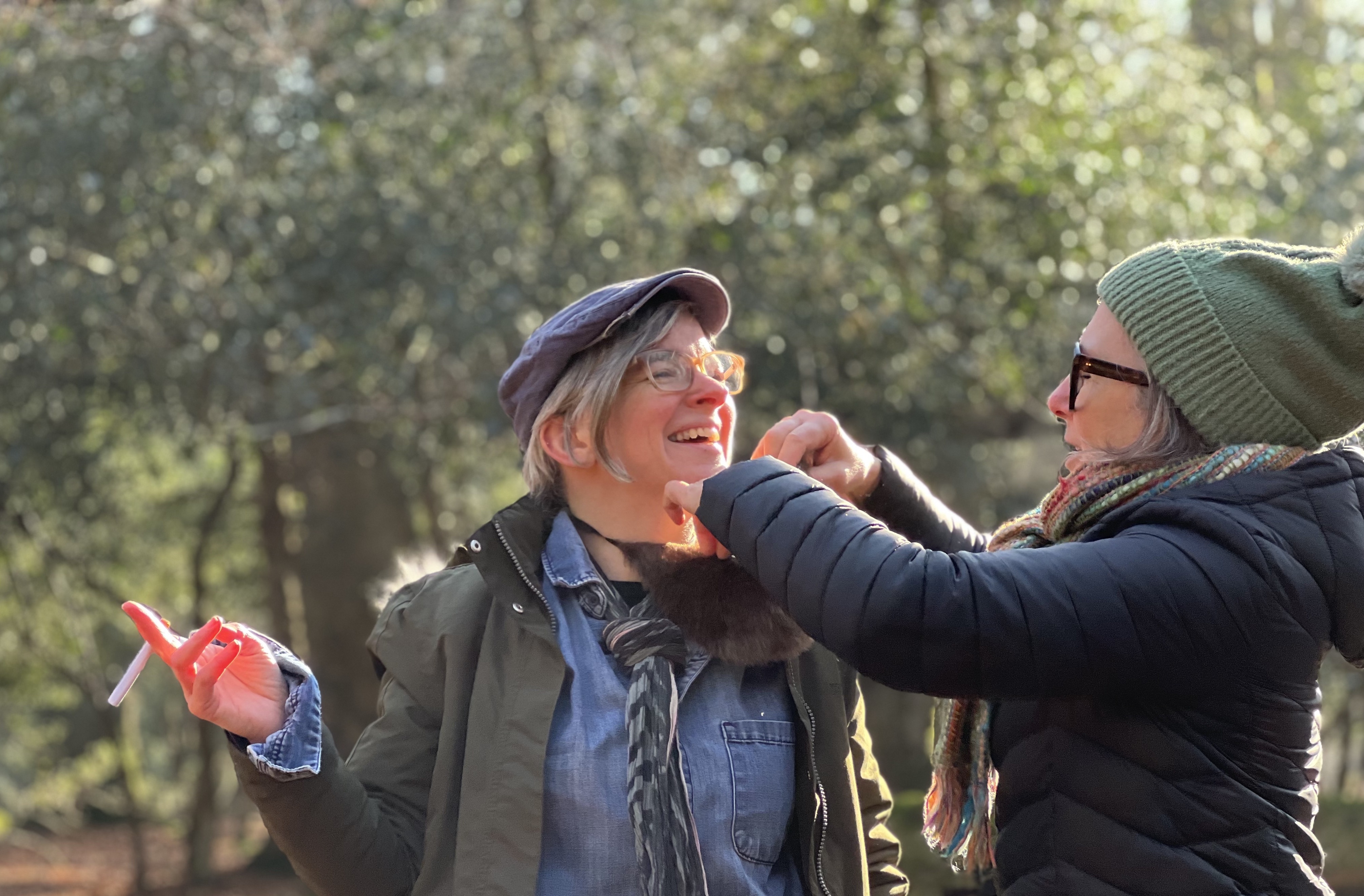 A person wearing a flat cap is smiling andlooking at another person wearing a woolly hat who is tying something to her neck.