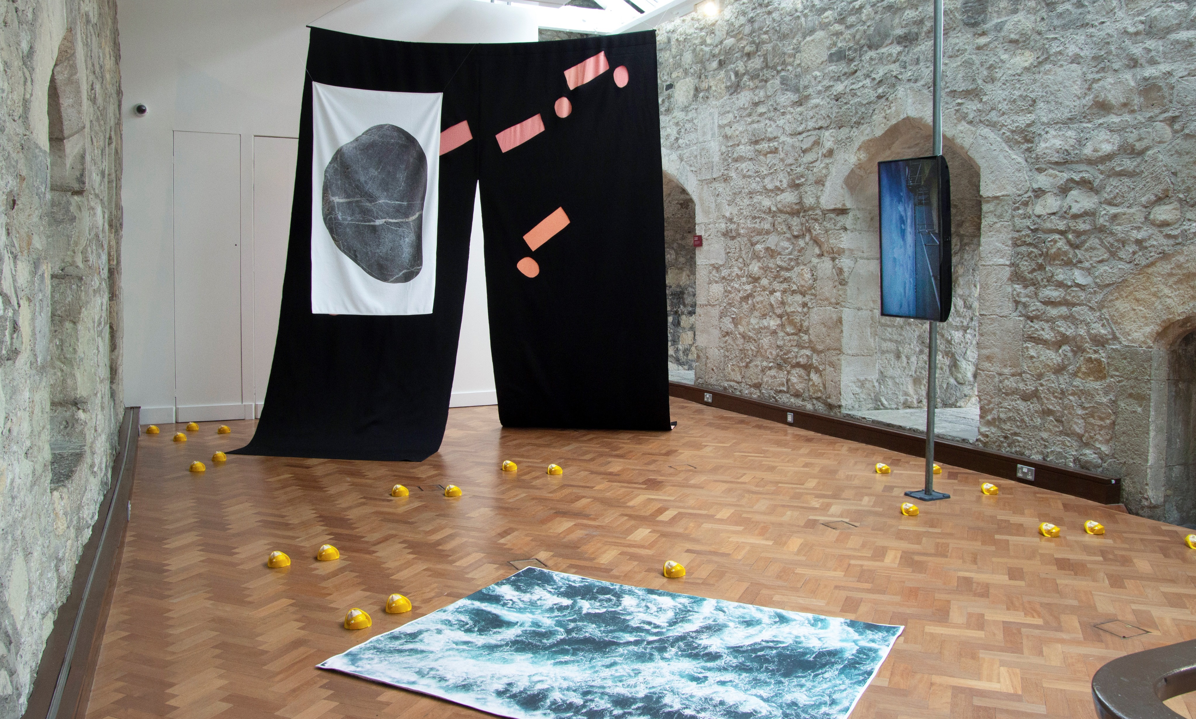 A gallery space with stone walls and windows and a wooden parquet floor. There is artwork installed including a film on a monitor strapped vertically to a metal pole; items scattered on the floor and a large hanging fabric work with a slit in the middle