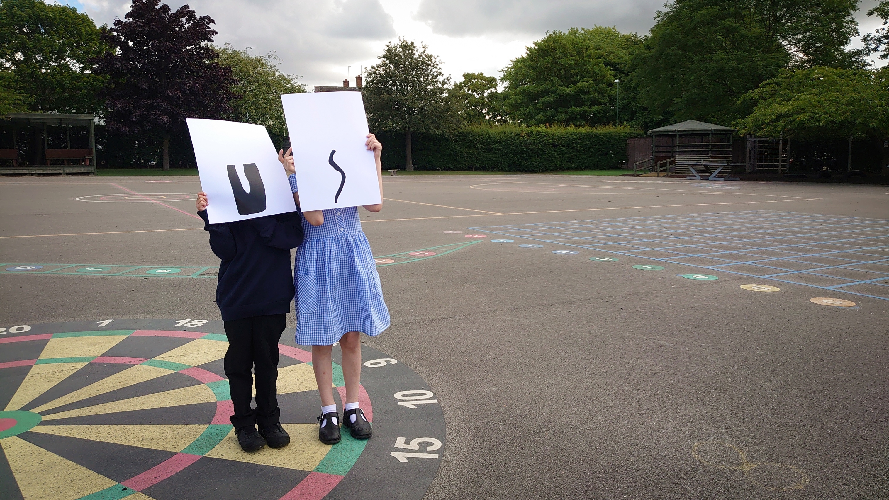 Two primary school children are stood in a school playground holding sheets of paper in front of their faces which have a single letter printed (U and S) on each sheet