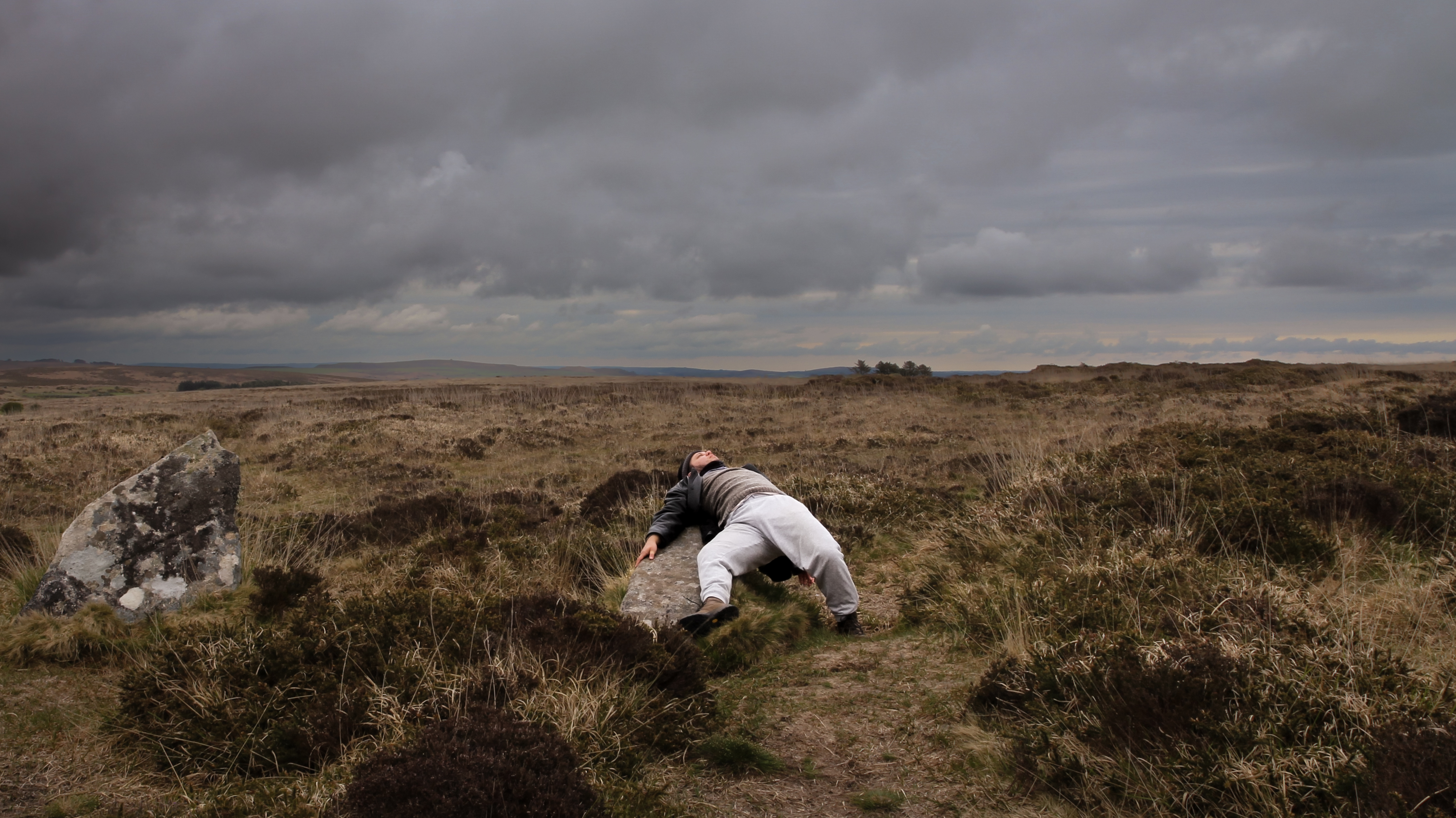 A person in a moor like, barren landscape, they are lying across a stone looking up to the sky