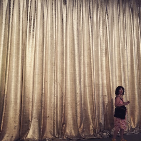 A woman walking past a large gold curtain looking to camera.