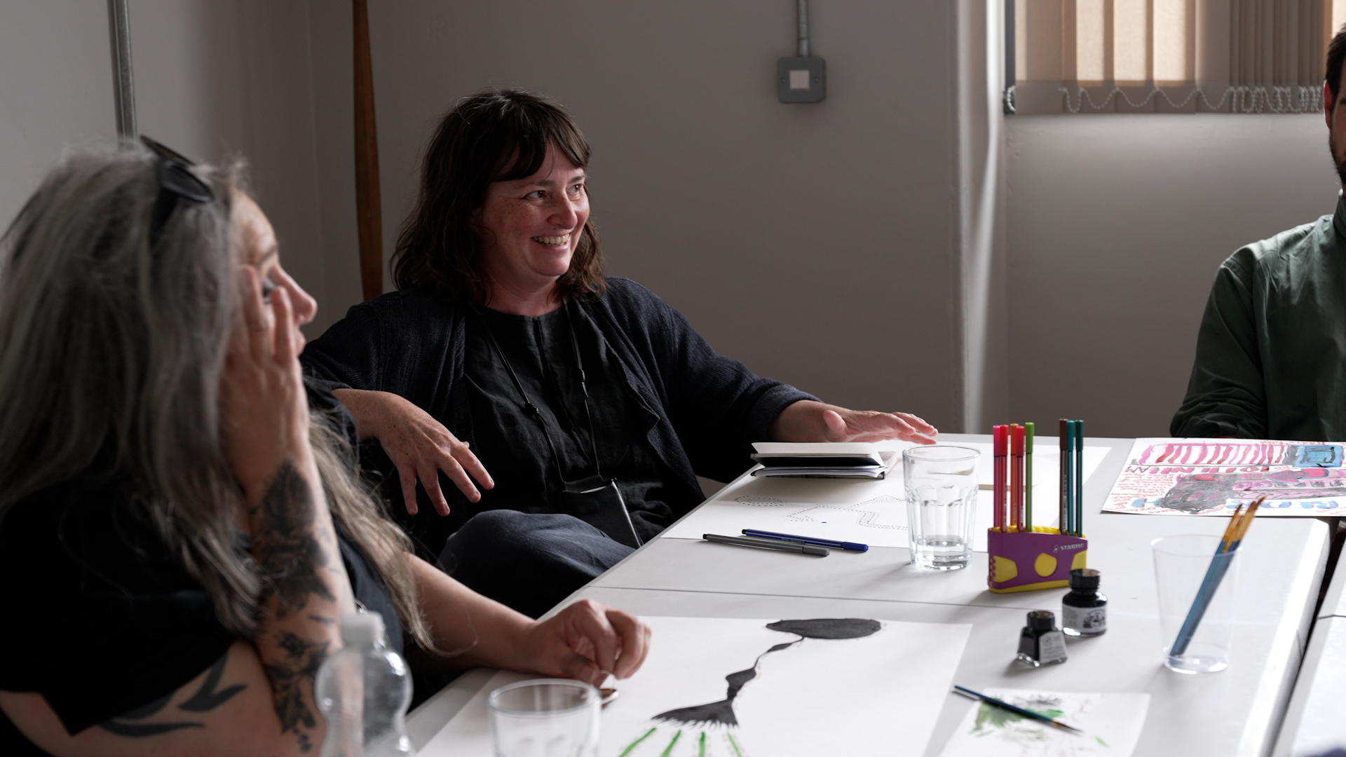 Two people sat next to each other around a table taking part in a discussion. One has long grey hair, their glasses propped on their head and their hand to their face. The other has shoulder length brown hair and is smiling, looking across to others sat at the table.