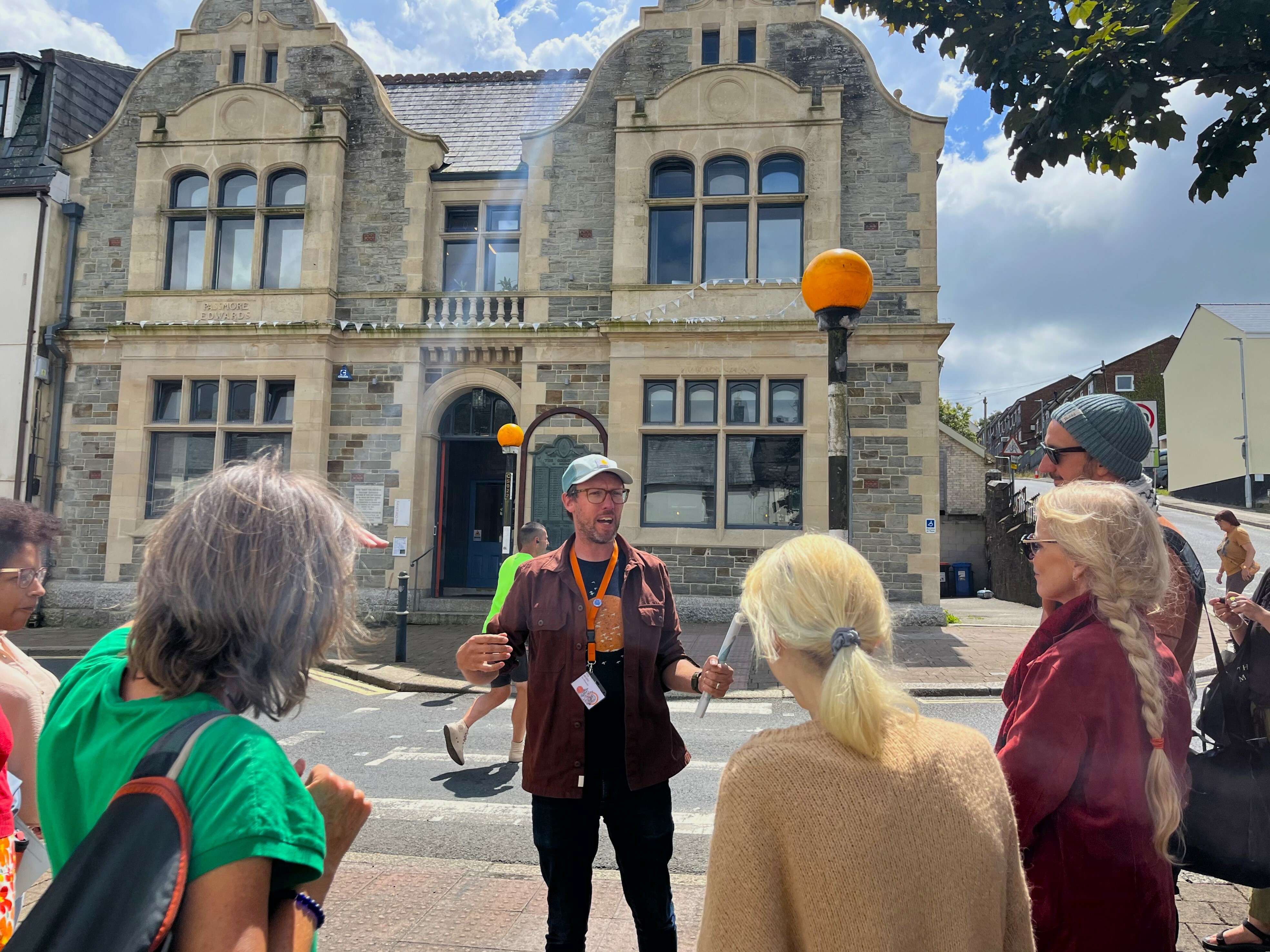 A man stood on a street with a stone building behind him is leading a guided tour. He is wearing an orange lanyard and speaking to a group gathered around.