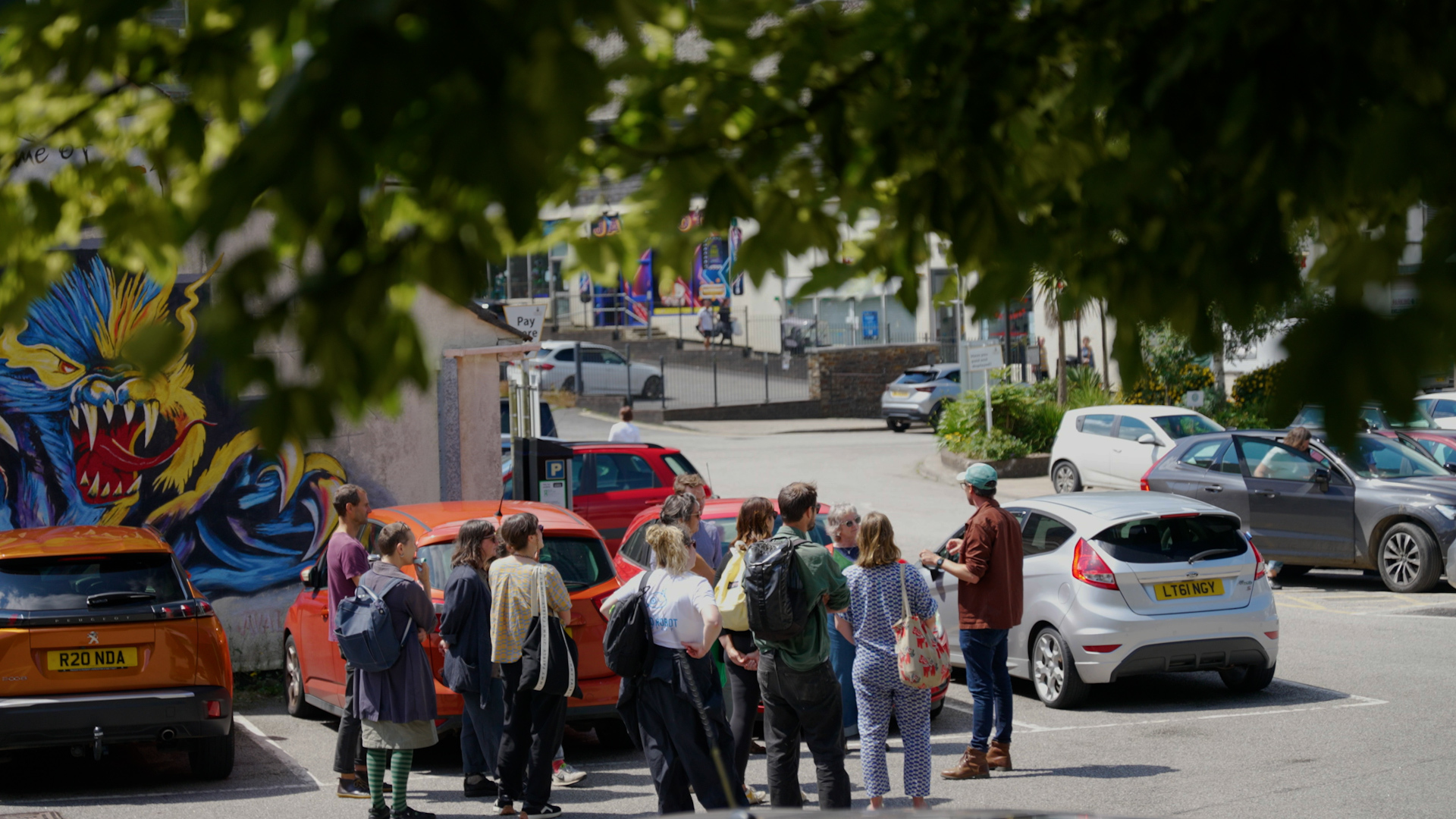 A group of 12 people are gathered in a carpark listening to a man at the front of the group who is leading a guided tour.