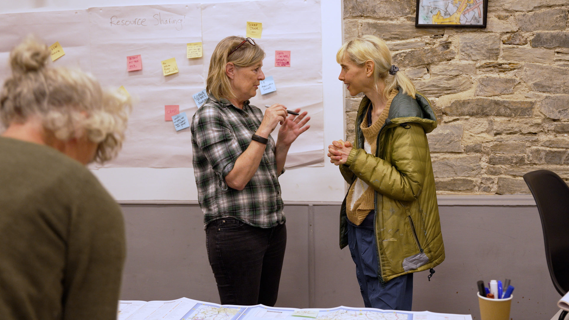 Two women are chatting at an event, one is wearing a checked shirt and has their glasses propped in their shoulder length hair. The other has their blonde hair tied in a ponytail and is wearing a green raincoat.