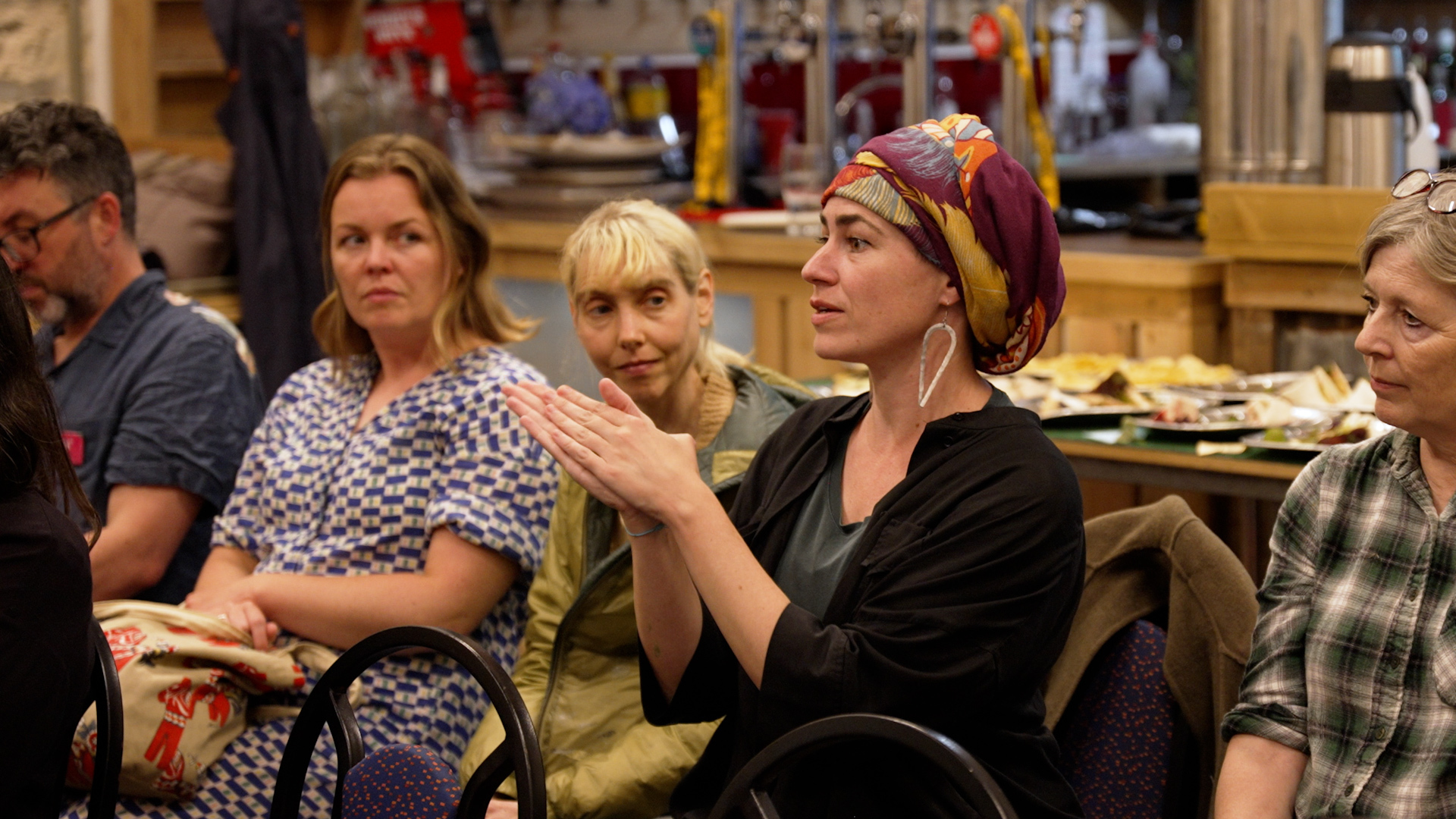 A woman sat next to others on a row of chairs is speaking to a roof of people, she has a fabric wrap covering her head and large silver earrings.