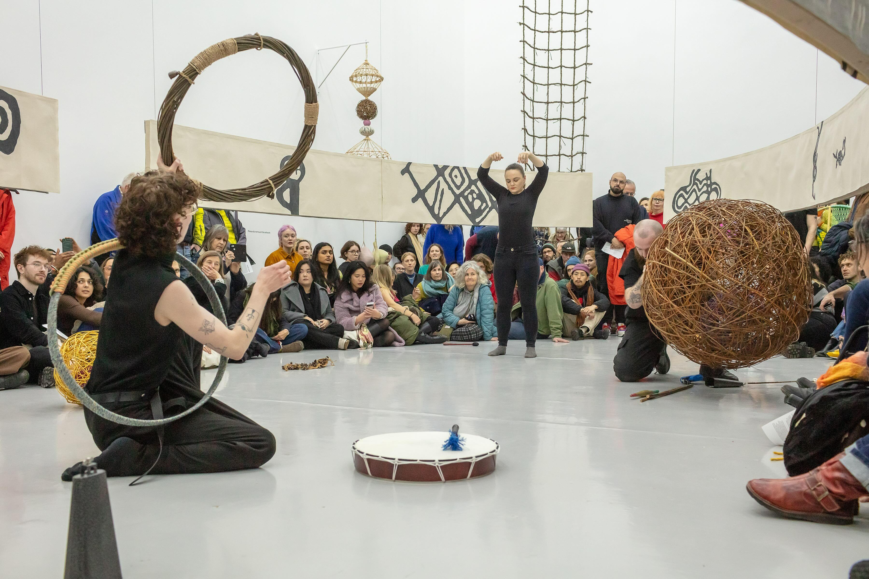 A photograph of an event that is part of an installation in a gallery space. The exhibition consists of sculptural objects including a large fabric banner that encircles the central area, a rope net structure that hangs from the ceiling and a wicker sculpture hanging from a wall. Three performers sit and stand in the central area, they are dressed in black and are activating a series of smaller objects and instruments.