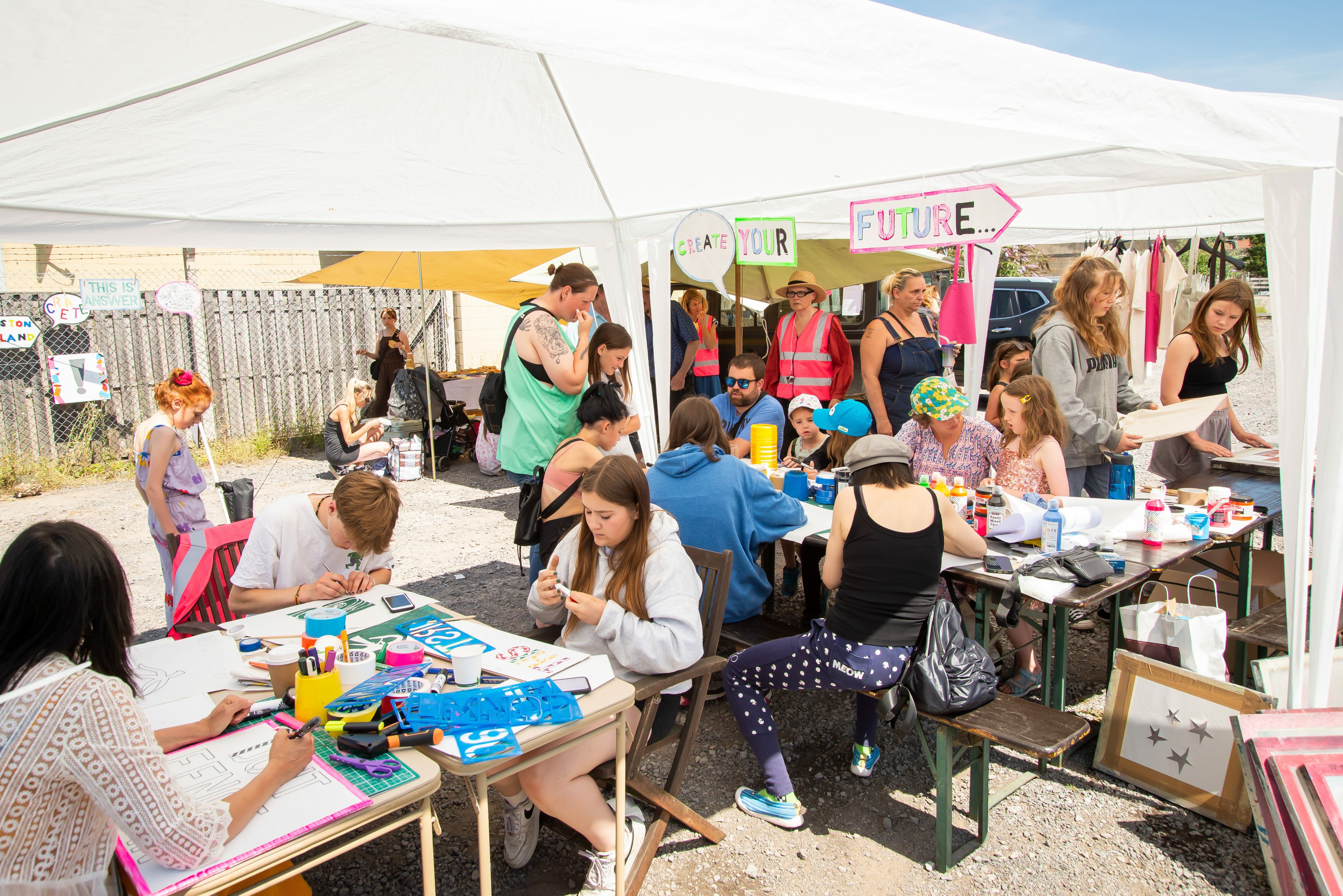 Several individuals seated beneath a white tent, engaged in drawing, colouring and stencilling. There are handmade signs in the background that say 'Create', 'your' and 'future'.