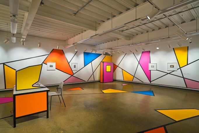 A gallery view of an installation consisting of colourful wall panels, framed works, and a desk with monitor