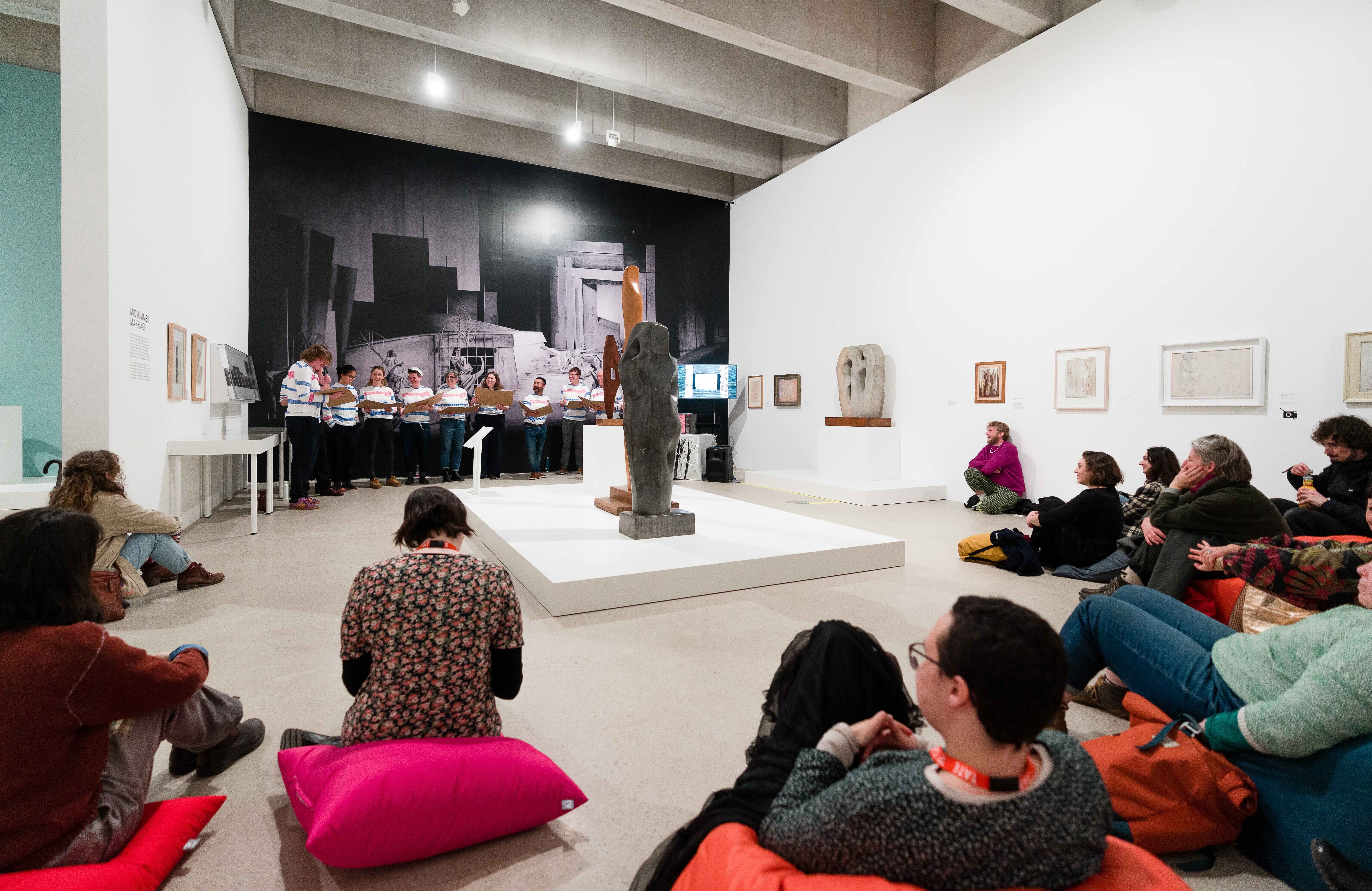 A photograph of an event in a gallery space. There is a choir at the far end of the gallery, they are standing in front of a large black and white photographic wall work. There are framed works on the walls to the left and right, and a plinth with sculptures in the middle. An audience is in the foreground, seated on colourful beanbags