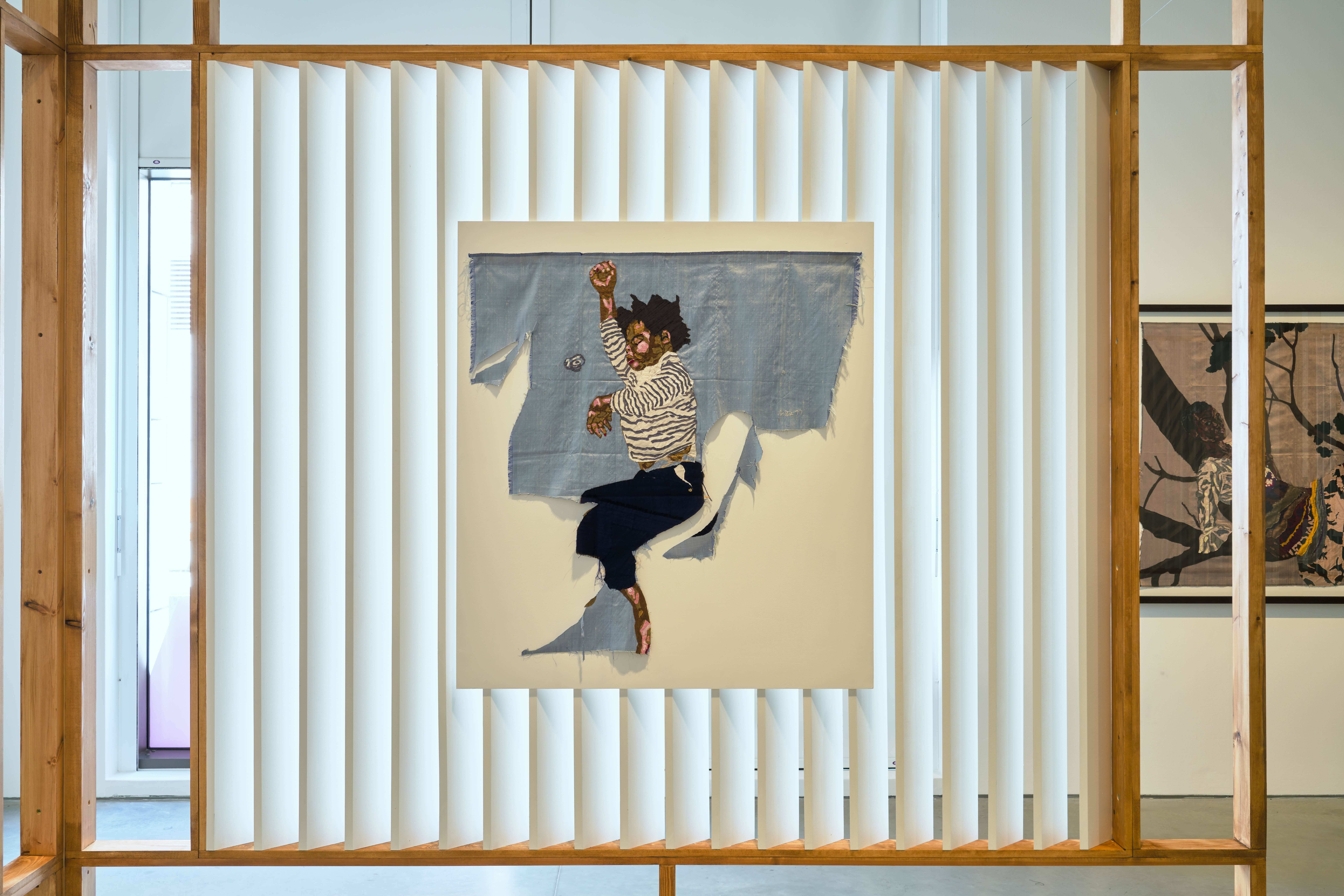 A photograph of an artwork consisting of a sleeping child stitched on to a blue silk panel. The panel is torn around the bottom of the figure, and attached to a white background. The work is presented in an installation made up of a series of long white vertical panels, mounted in a wooden frame