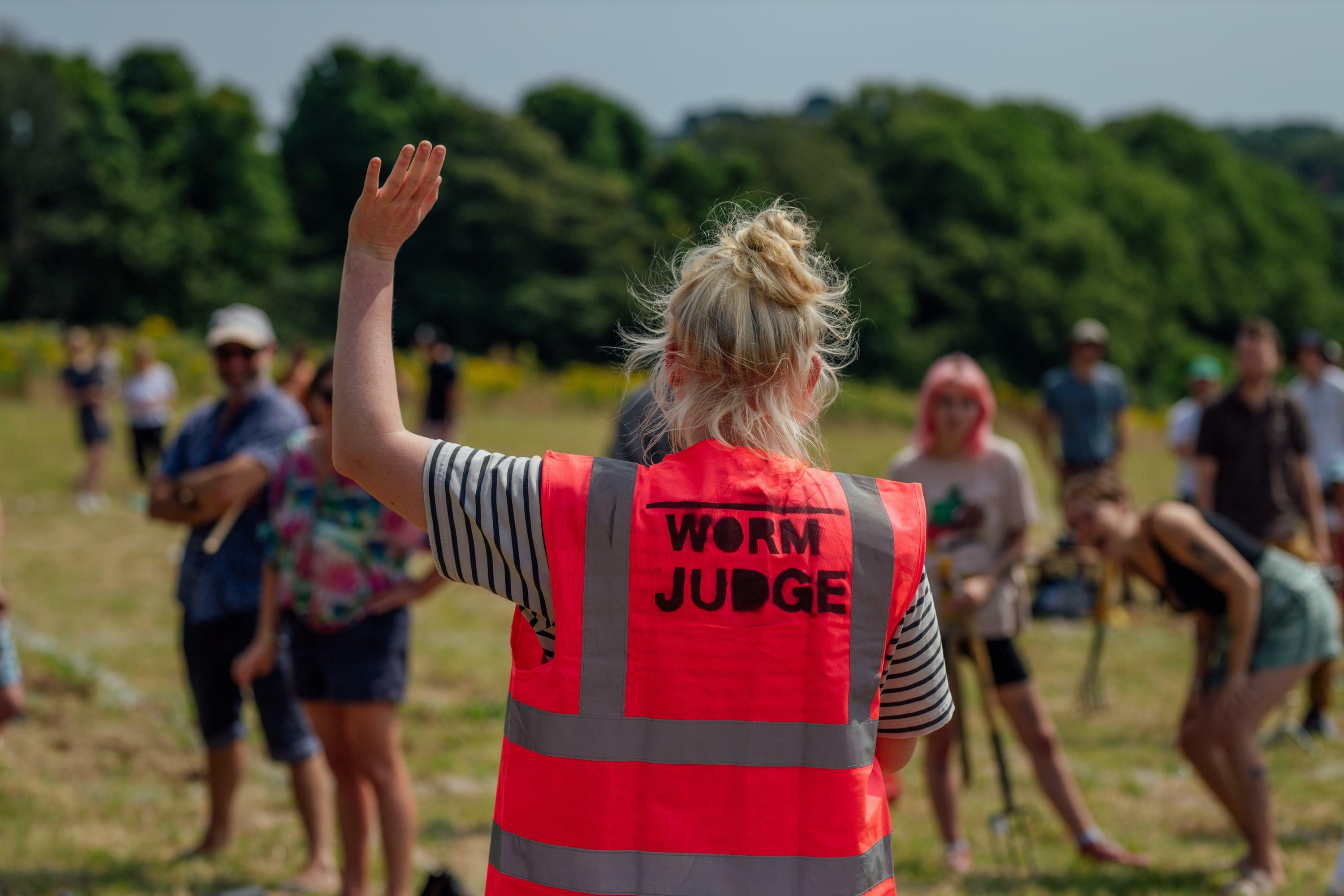 A person wearing a pink h-vis jacket with 'Worm Judge' printed on the back, have their back to the camera and hand in the air talking to people gathered round.