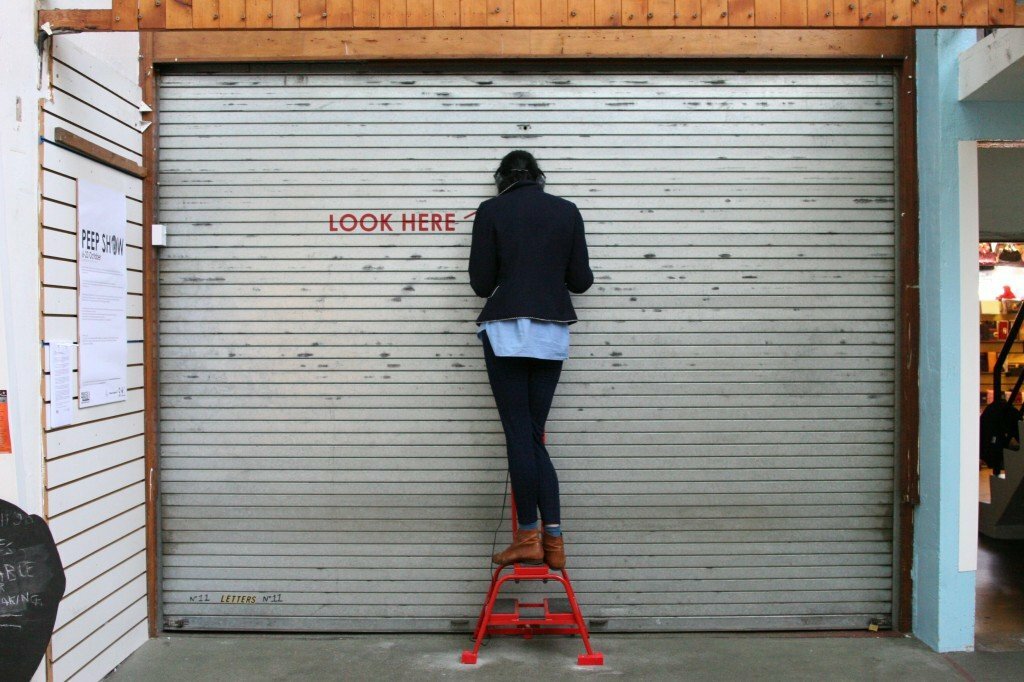 A person stood on a red step stall looks into a peep hole located on a shop shutter. The shutter has the words "LOOK HERE" and an arrow printed on it.