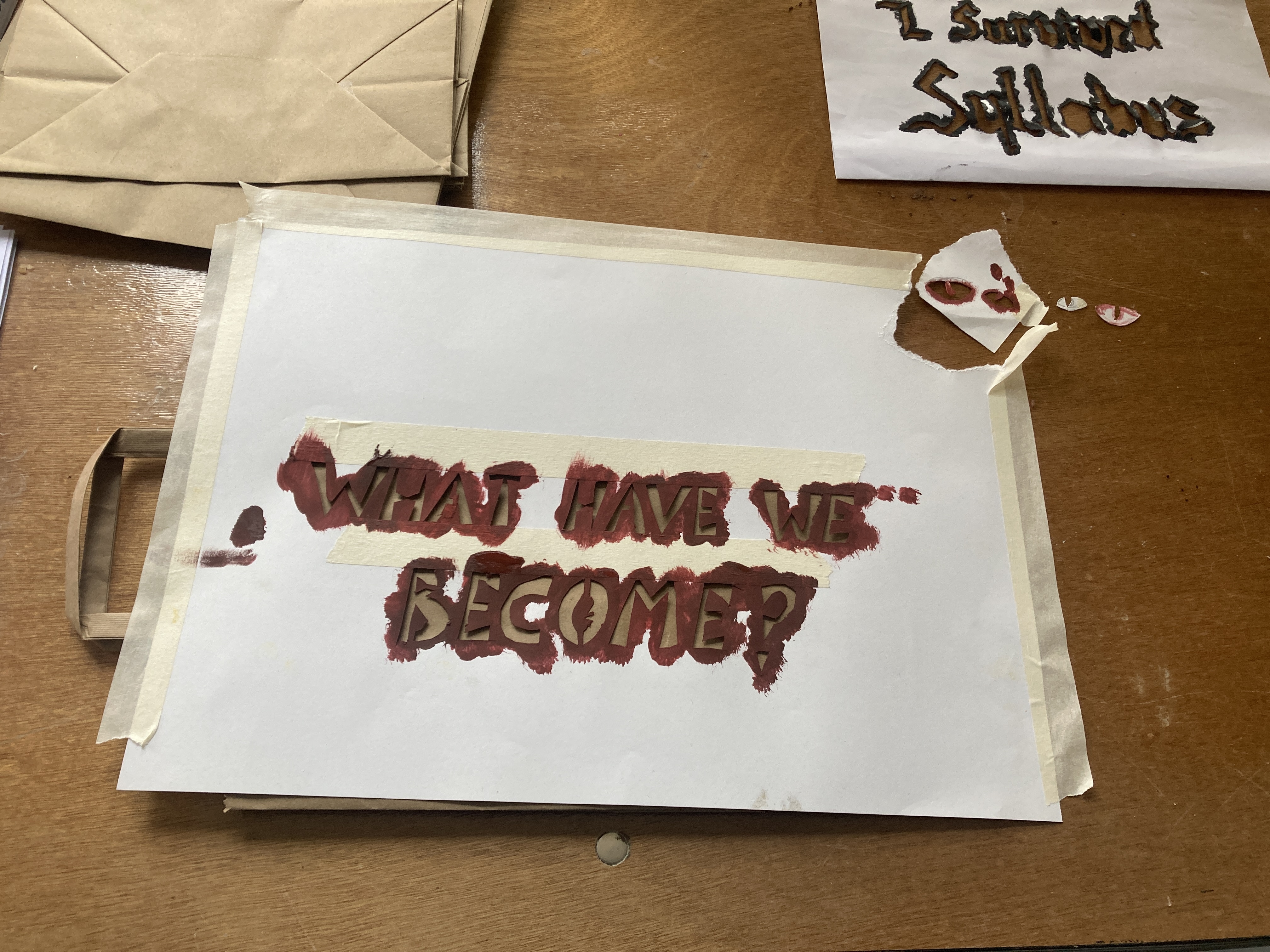 A photograph of a paper stencil with the words "what have we become" cut out. The stencil has been used, and there is masking tape around the edges, dark red paint around the letters, and the top right corder has been torn off.