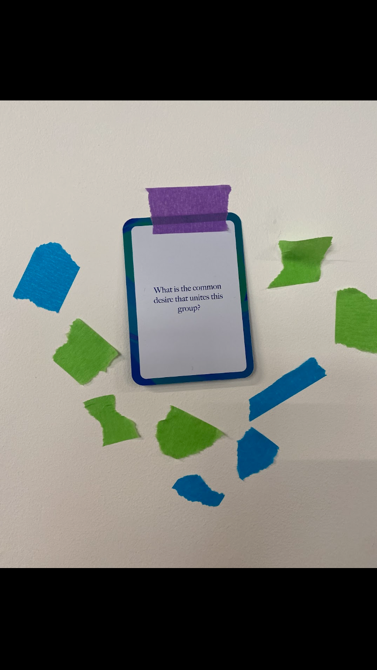 A photograph of a card taped to a wall with purple masking tape with the words "What is the common desire that unites this group. There are other pieces of green and blue masking tape surrounding the card.