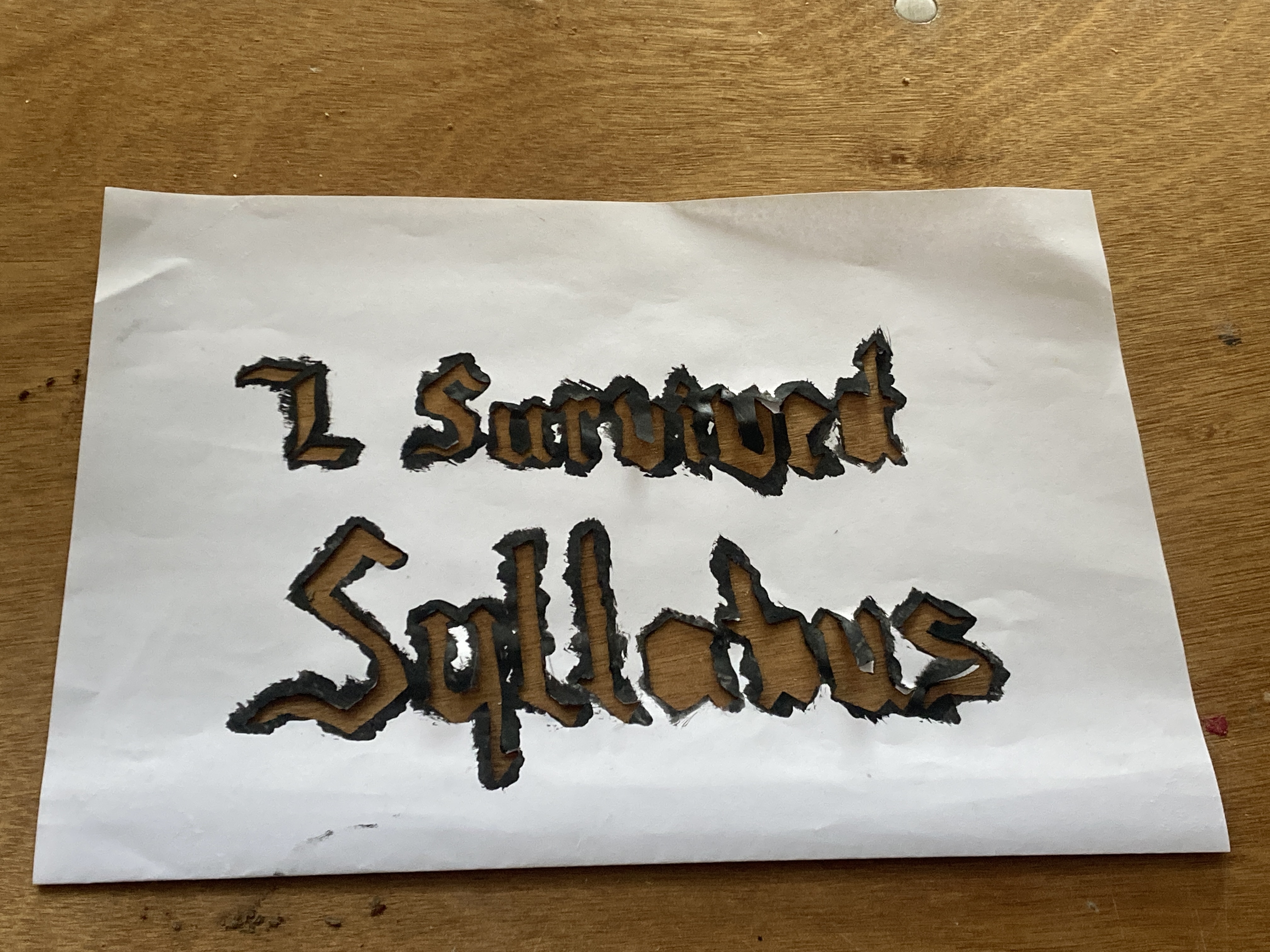 A photograph of a paper stencil with the words "I survived Syllabus" cut out. The stencil is on a wooden table top.