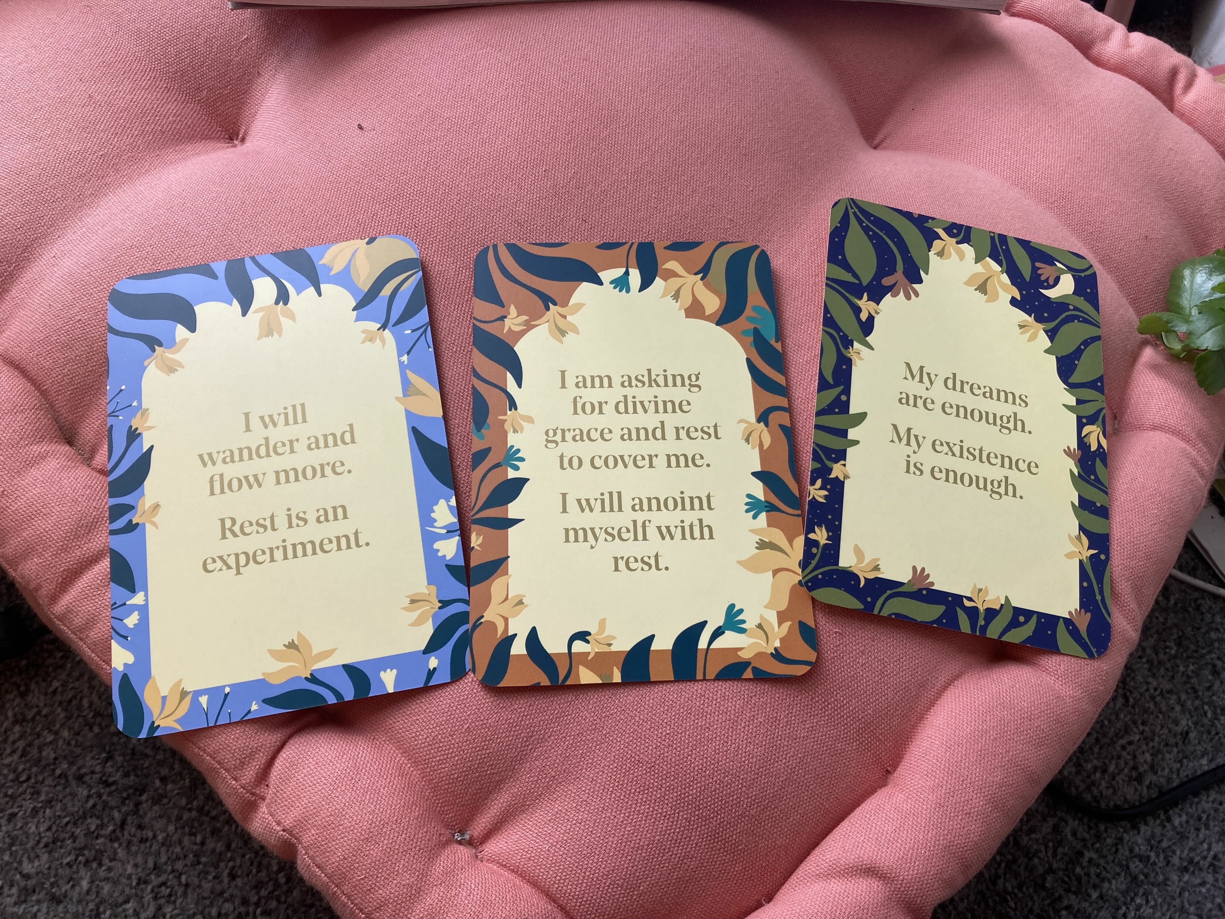 A photograph of three cards on a bright pink cushion. Each card has a short text relating to rest, and is illustrated around the edges with plants and flowers.