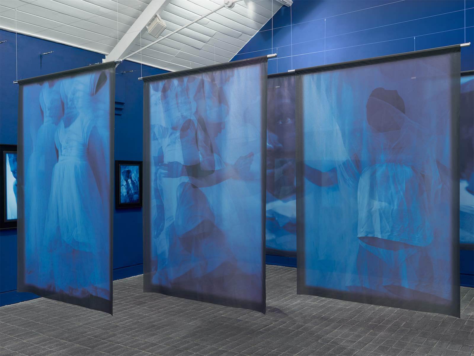 A series of hanging blue tinted banners with images of figures wearing white clothing printed on them in a blue walled gallery space