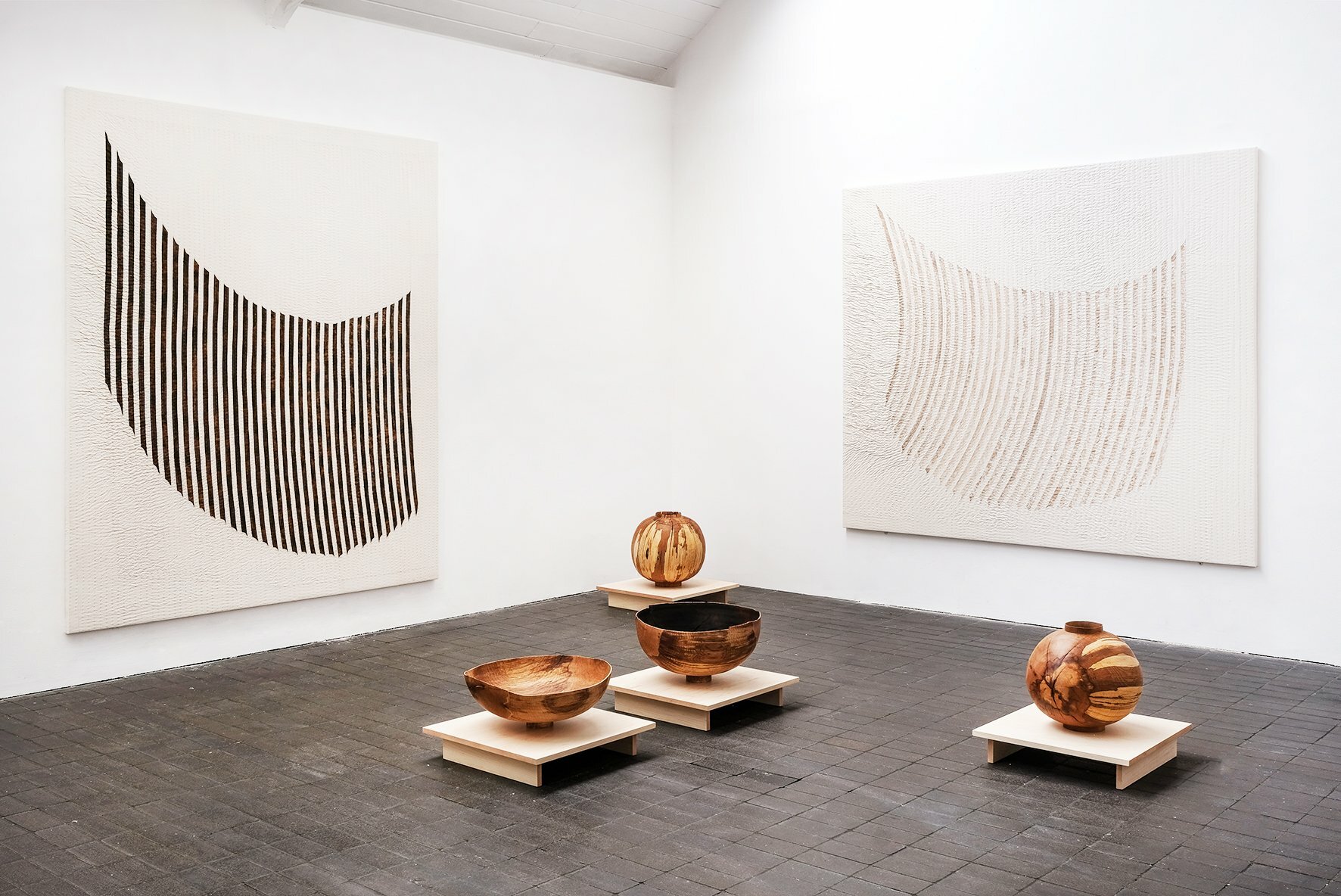 Two large monotone paintings hanging on a gallery wall with large hand carved wooden bowls placed on plinths on the floor in front of the paintings