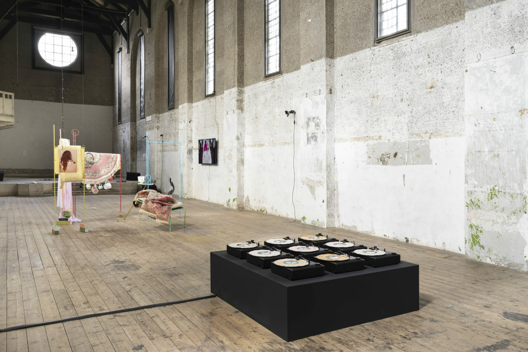 A grid of nine turntables set up on a black plinth as part of a group exhibition in a large empty church. There is a sculptural installation in the background with colourful metal framework with carpets draped over.