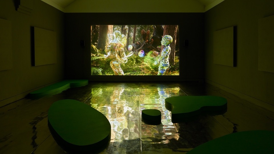 A gallery room with green walls and bathed in green light. There are large shapes on the floor made in wood and painted green which resemble Lilly pads. A large video p[projection is on the wall showing digital figures in a computer generated forest environment.