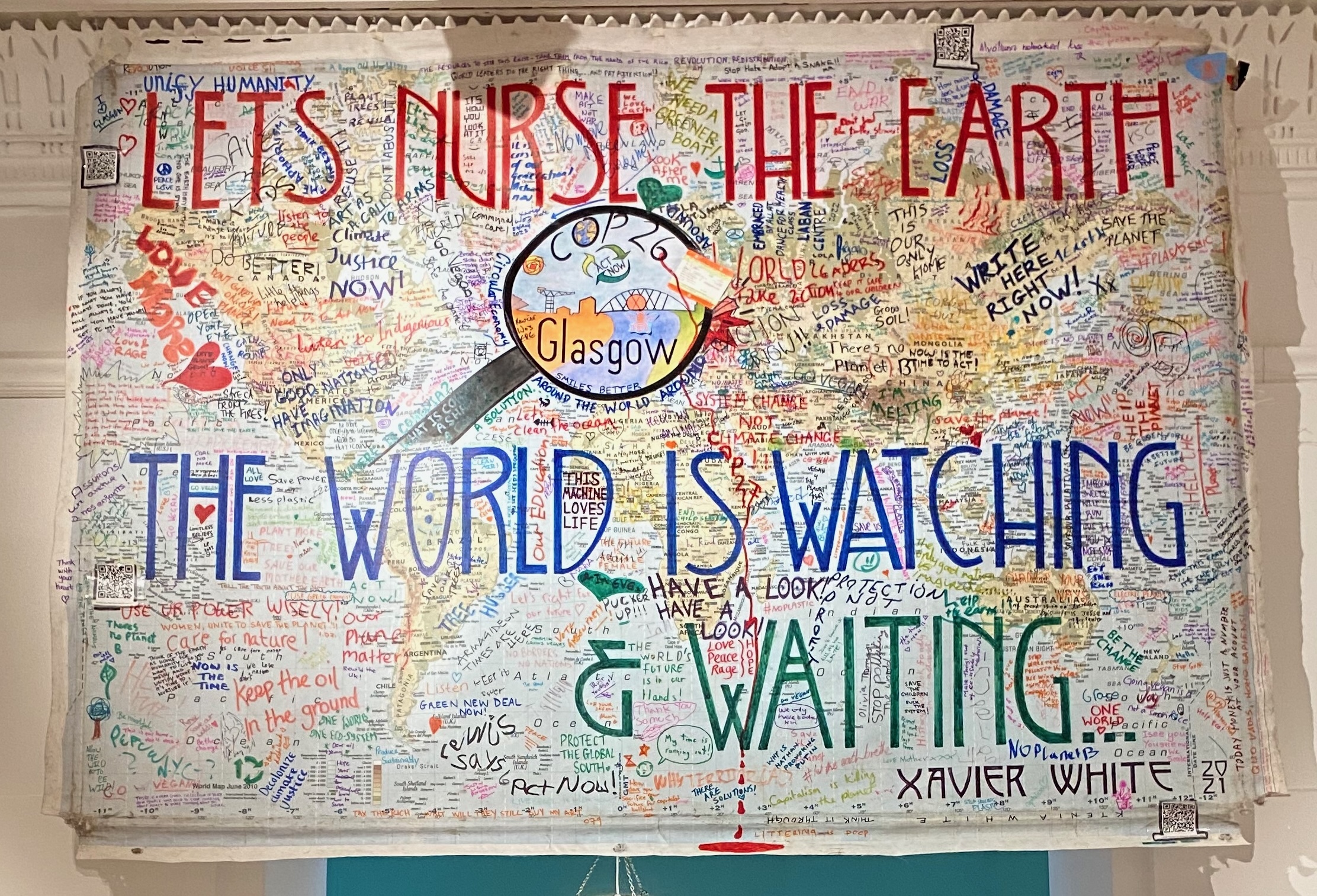 An artwork consisting of a large map which has been overwritten with text, including 'Let's Nurse the Earth, the World is Watching' in large letters. There is an illustration of a magnifying glass with with words 'Glasgow COP 26' in the centre