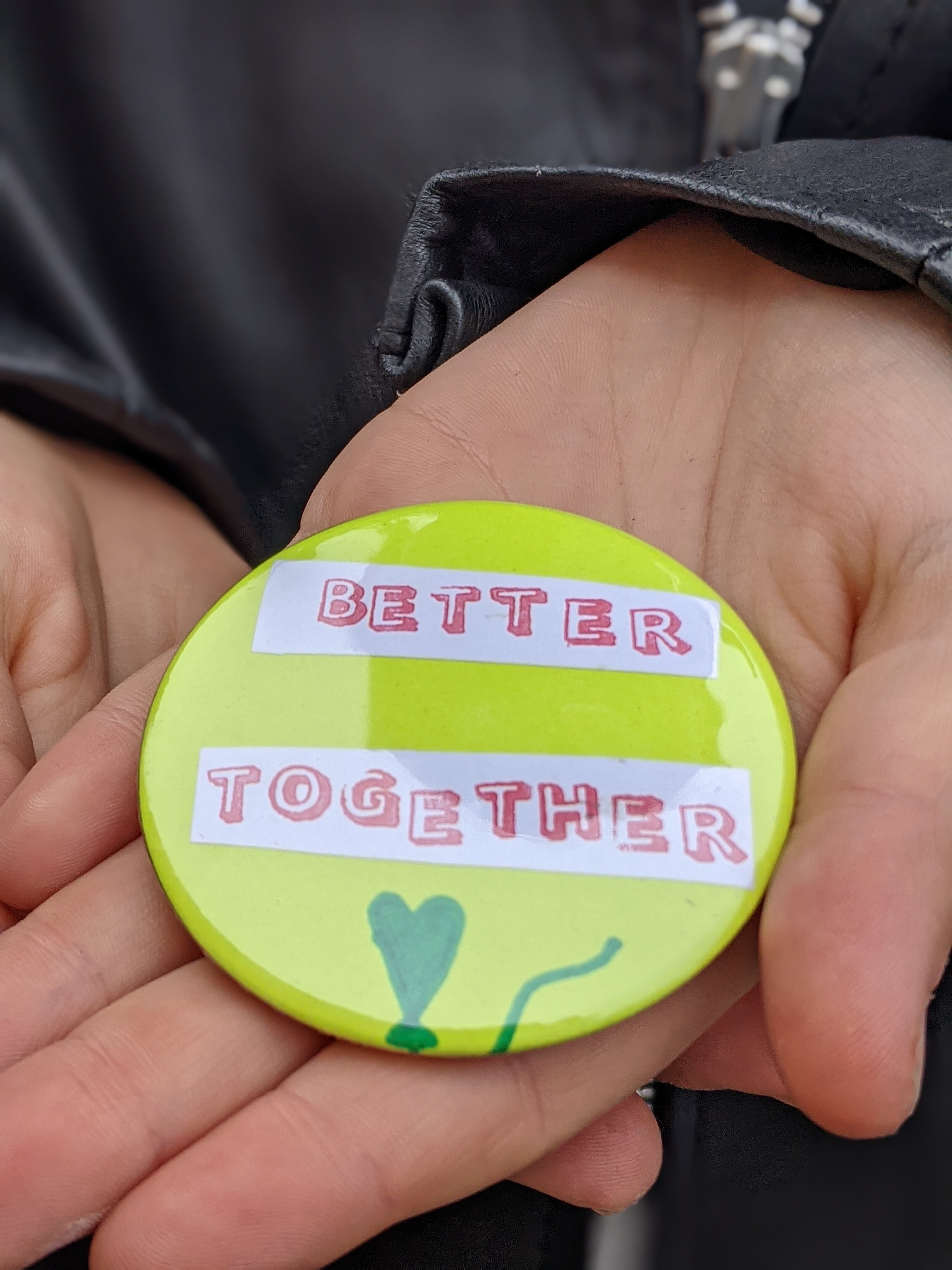 Person holding a lime green badge in the palm of their hand. The badge has the text 'better together' and a drawing of a heart on it with sticker.