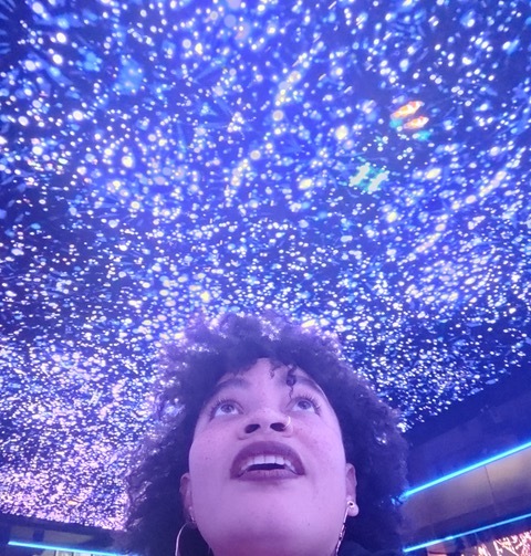 A portrait of a black woman shot from below looking up to a purple-lit ceiling covered in small lights