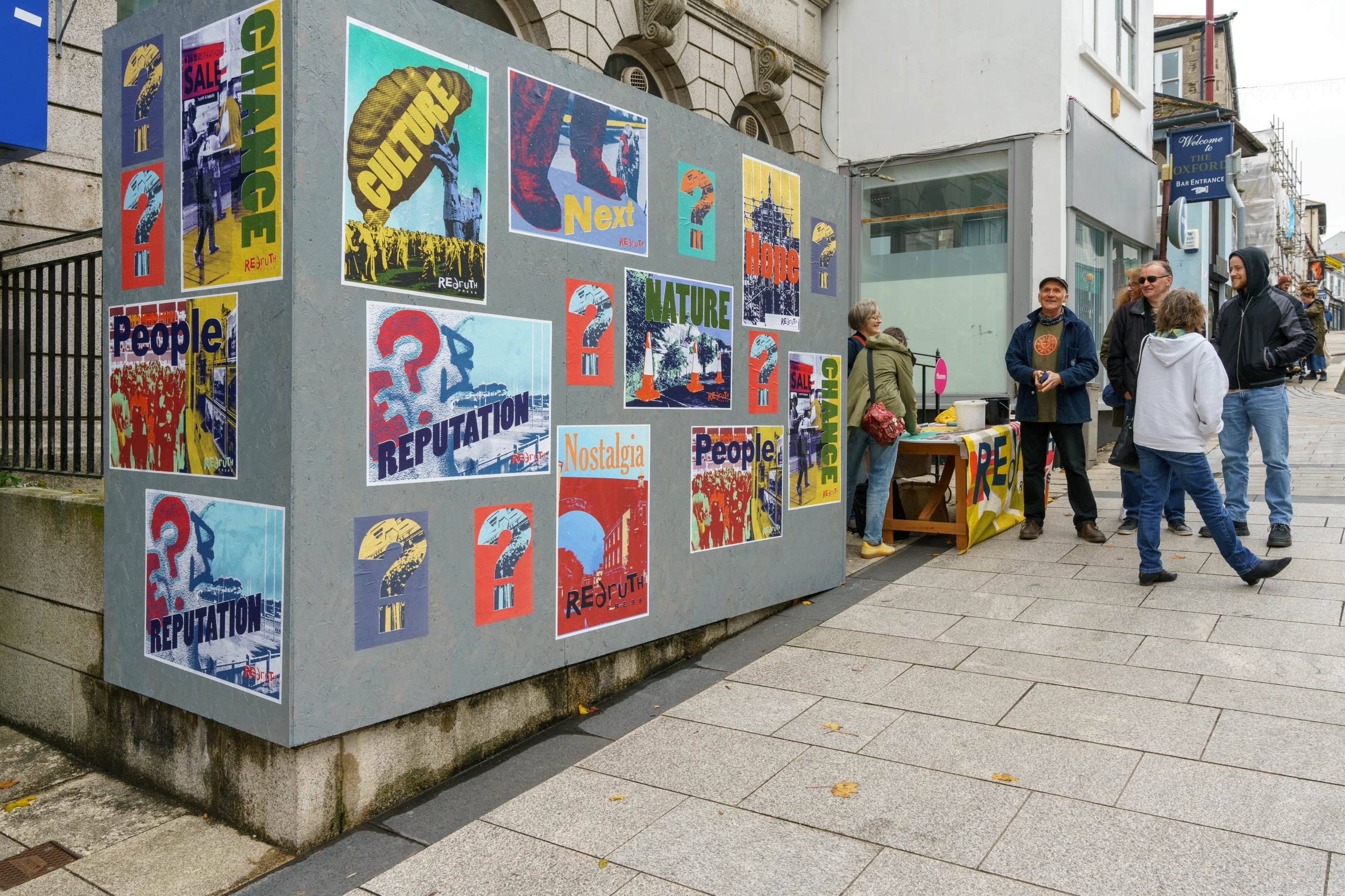 A set of hoardings painted grey wrap around the front of a building on a high street. There are graphic posters flyposted on the hoardings with texts that say things like 'culture', 'reputation', 'people', and 'nature'. There is a stall set up next to the hoardings and a group of people chatting