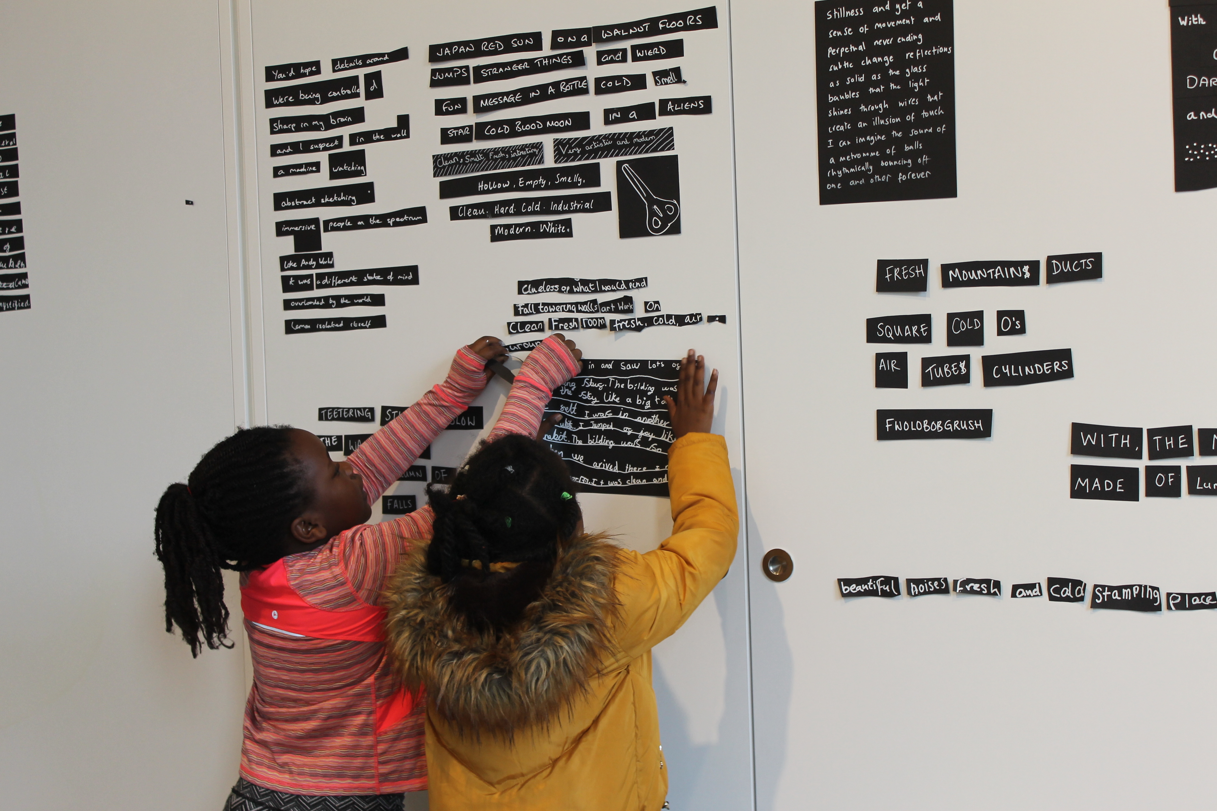Two young children have their backs to the camera sticking up pieces of black paper with white writing on them to a wall. On the wall we can see lots of other pieces of paper already stuck up.