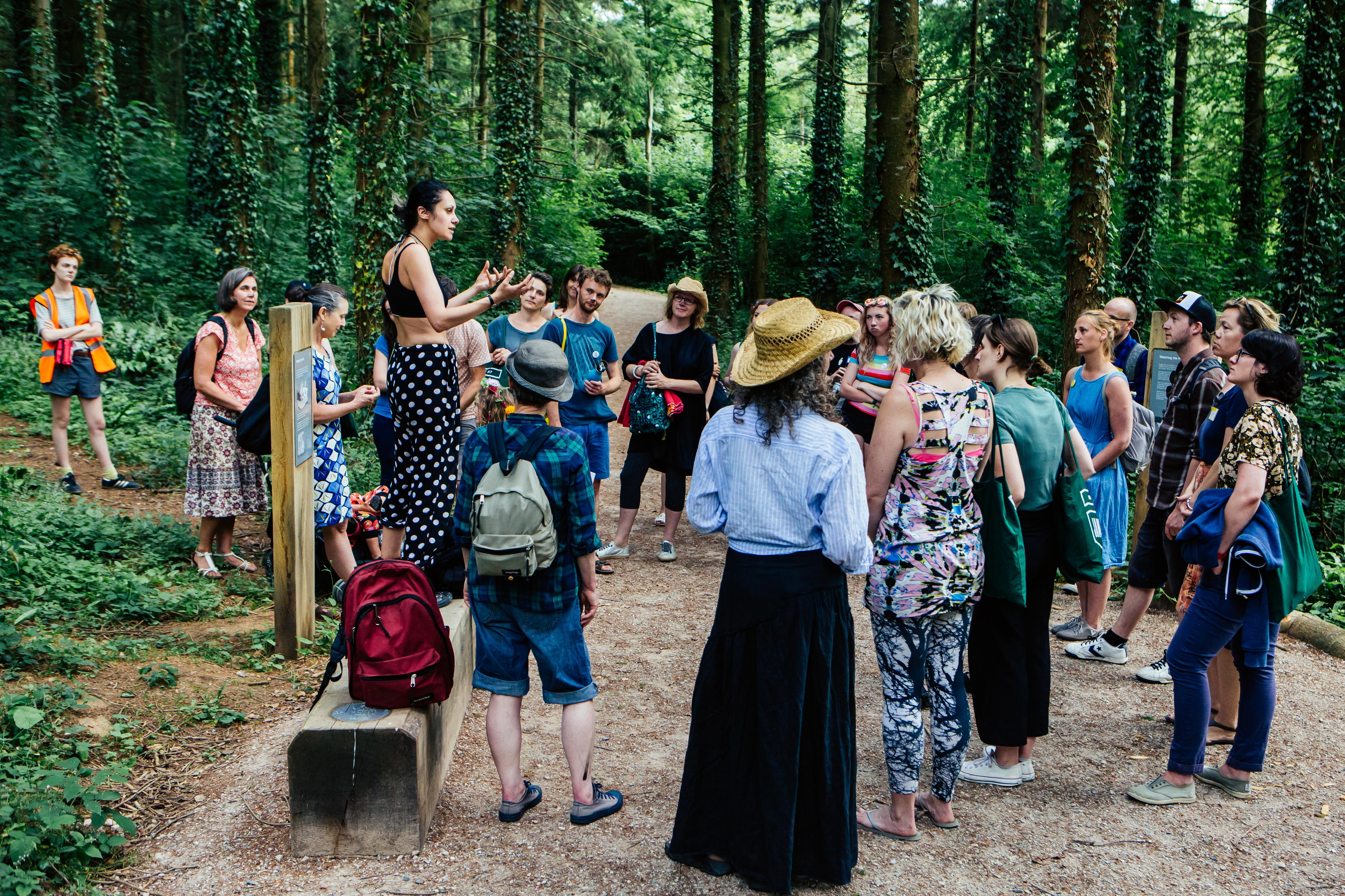 A group of people are stood on a path within a woodland, they are gathered around on a pathway listening to a woman who is stood on concrete bench speaking to them.