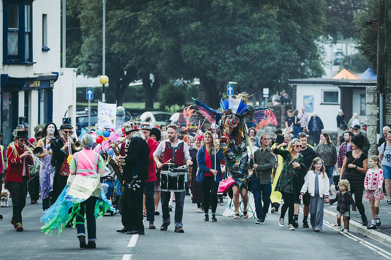 A festival-like parade of people walking down a road that has been closed to traffic. There are people carrying and playing instruments, a sea creature sculpture being carried and a a sign that says ‘This Land’