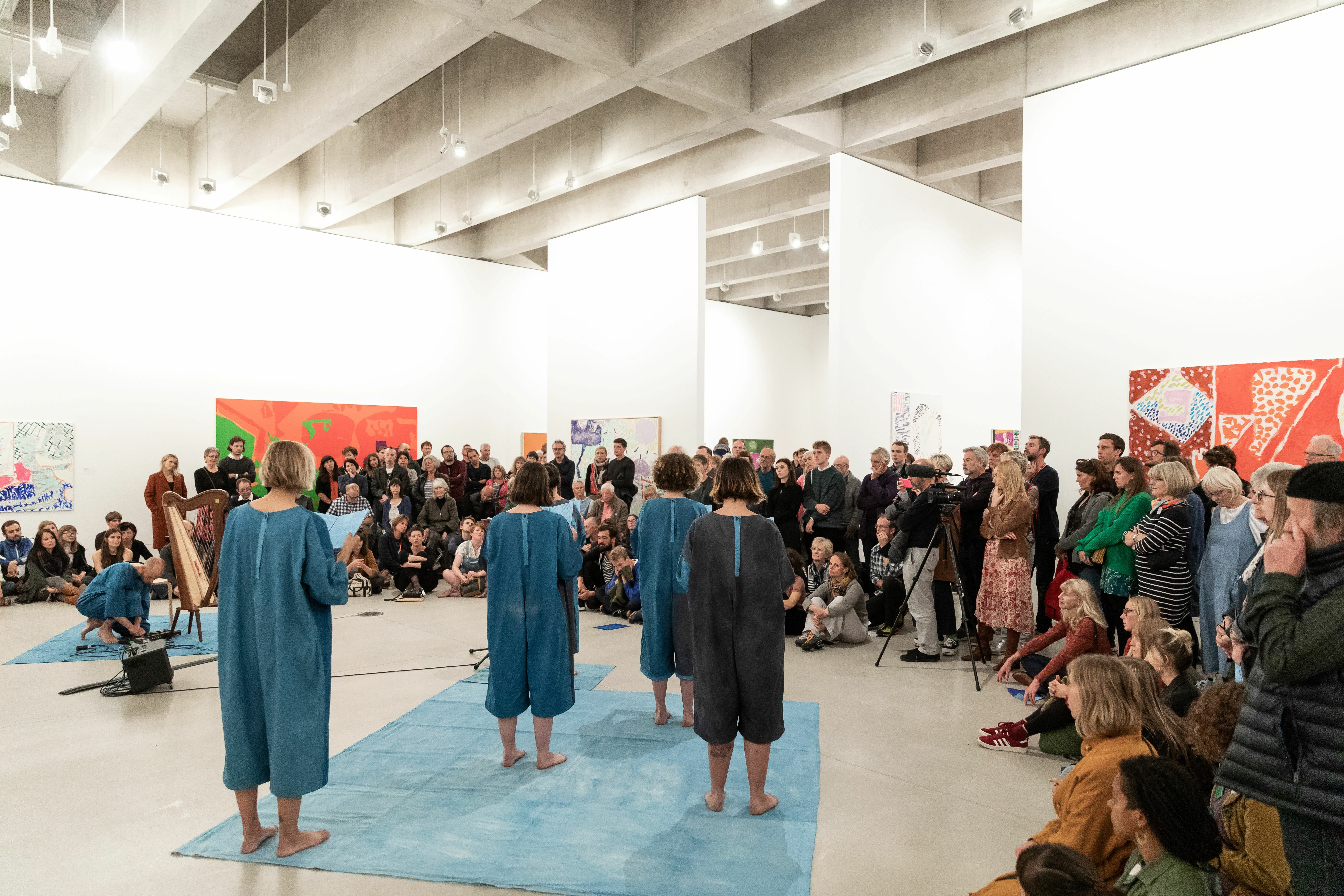 A group of people dressed in blue and grey onesies have their backs to the camera. They are stood on a blue fabric mat holding song booklets facing a crowd of people in a gallery space. To the left a person is crouching, adjusting knobs of a music mixer next to a harp
