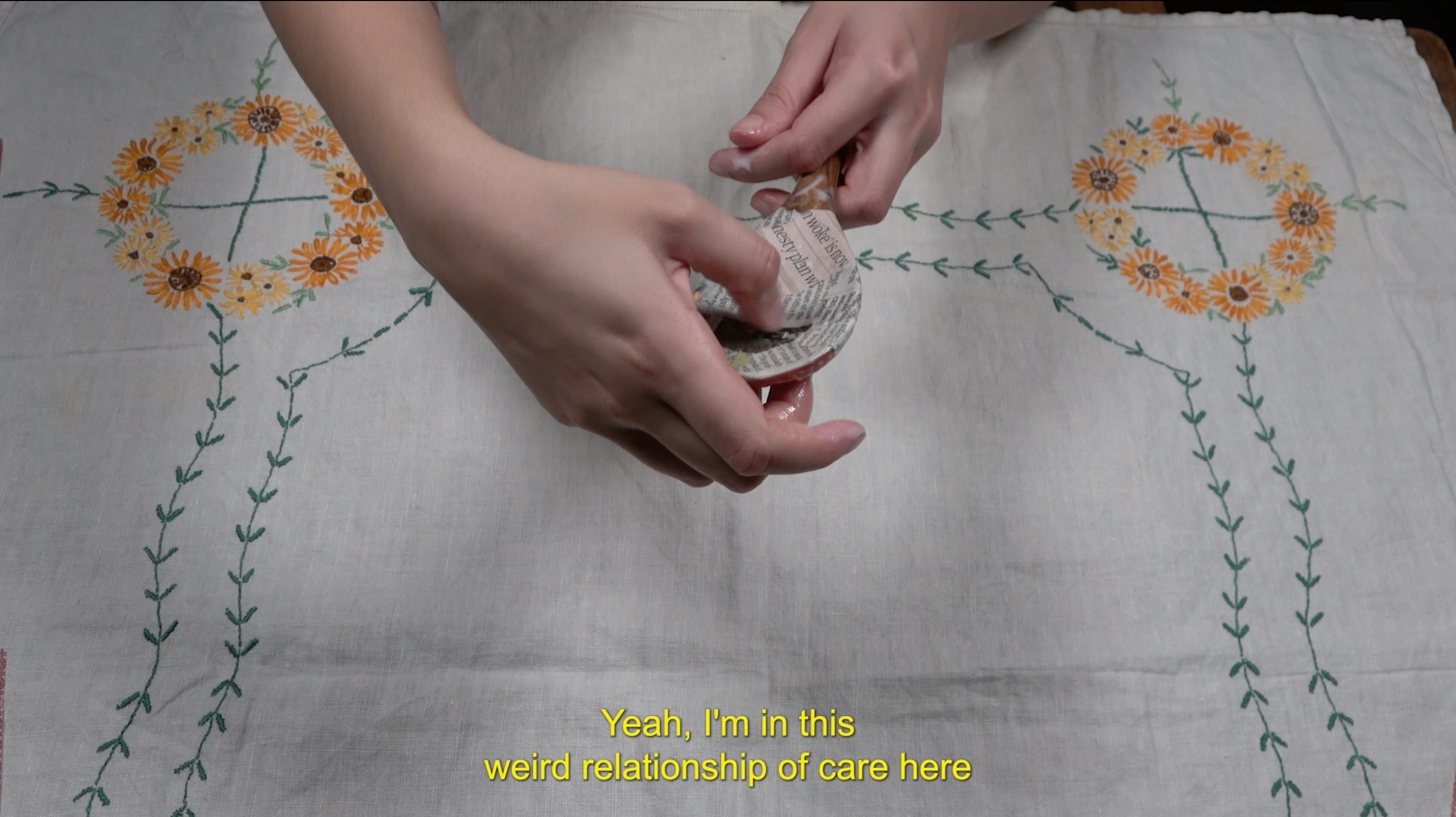 A film still showing two hands holding a spoon that has been covered in newspaper, over a white tablecloth embroidered with yellow and orange flowers. There are yellow subtitles at the bottom of the screen which say 'Yeah, I'm in this weird relationship of care here'