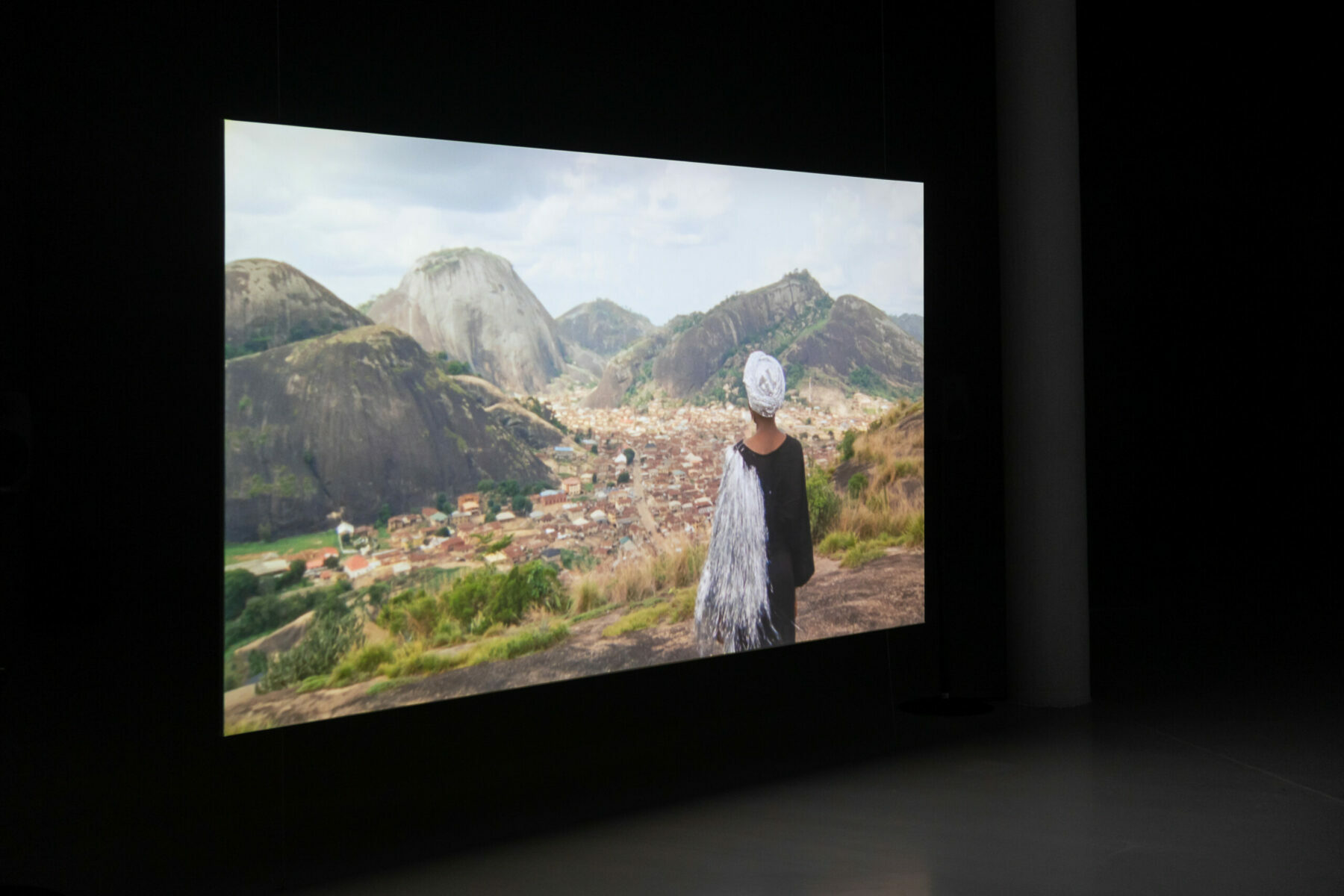 A video projection in a dark space, which shows the back view of a figure looking out onto a landscape.