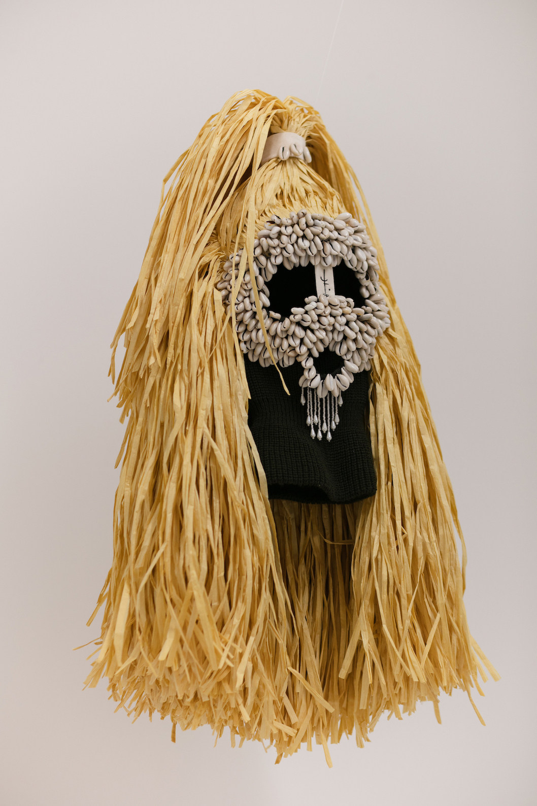 Image shows a mask with dark eyes with a face made of cowrie shells and surrounded raffia grass, between it's eyes is a symbol of a spell. The mask has a powerful gaze.