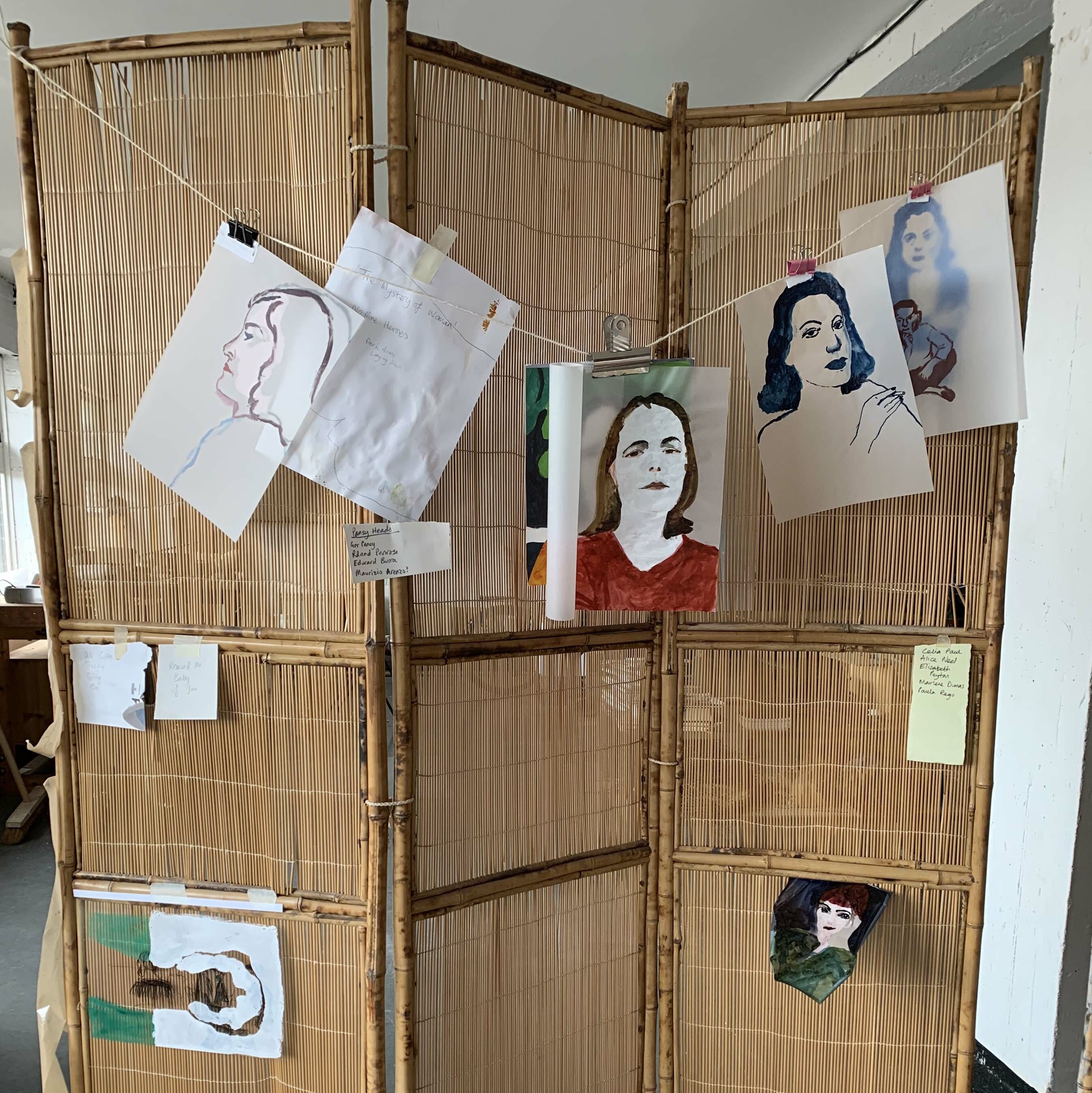 Several painted portraits on paper hang from a string line on an old wicker room divider