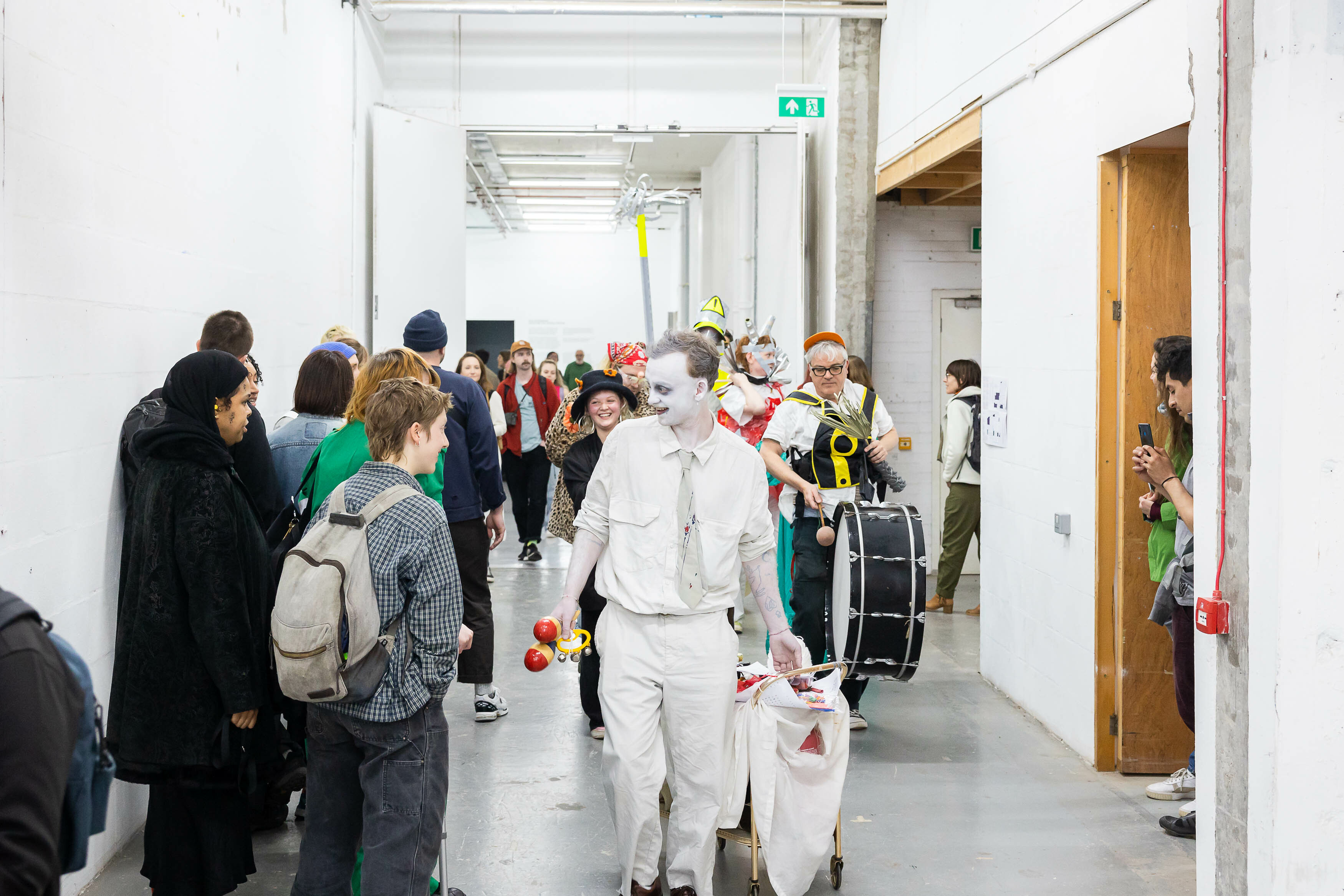 A procession of people, watched by a line of people. Performers holding instruments lead the procession through the hallways of Spike Island. The walls are white, with grey floors.