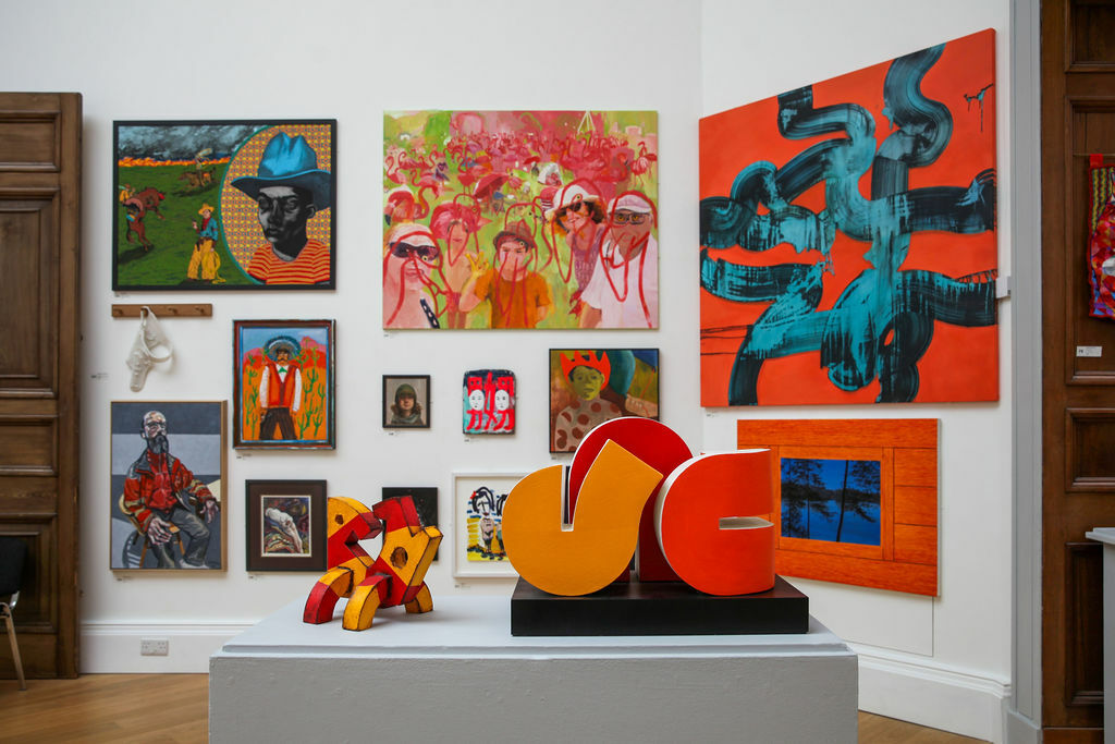 Photograph of the RWA Main Galleries with two red and yellow sculptures in the foreground and a full wall of colourful artworks in the background.