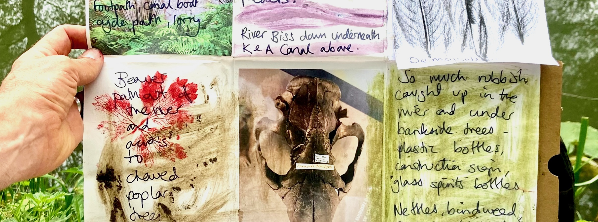 Outside in a woodland. Hand holding folded sketchbook page with handwritten text, leaf rubbings and collage.