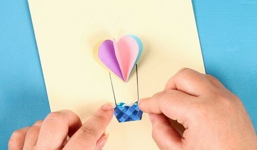 A pair of hands glue coloured card together to make a two-dimensional hot air balloon.