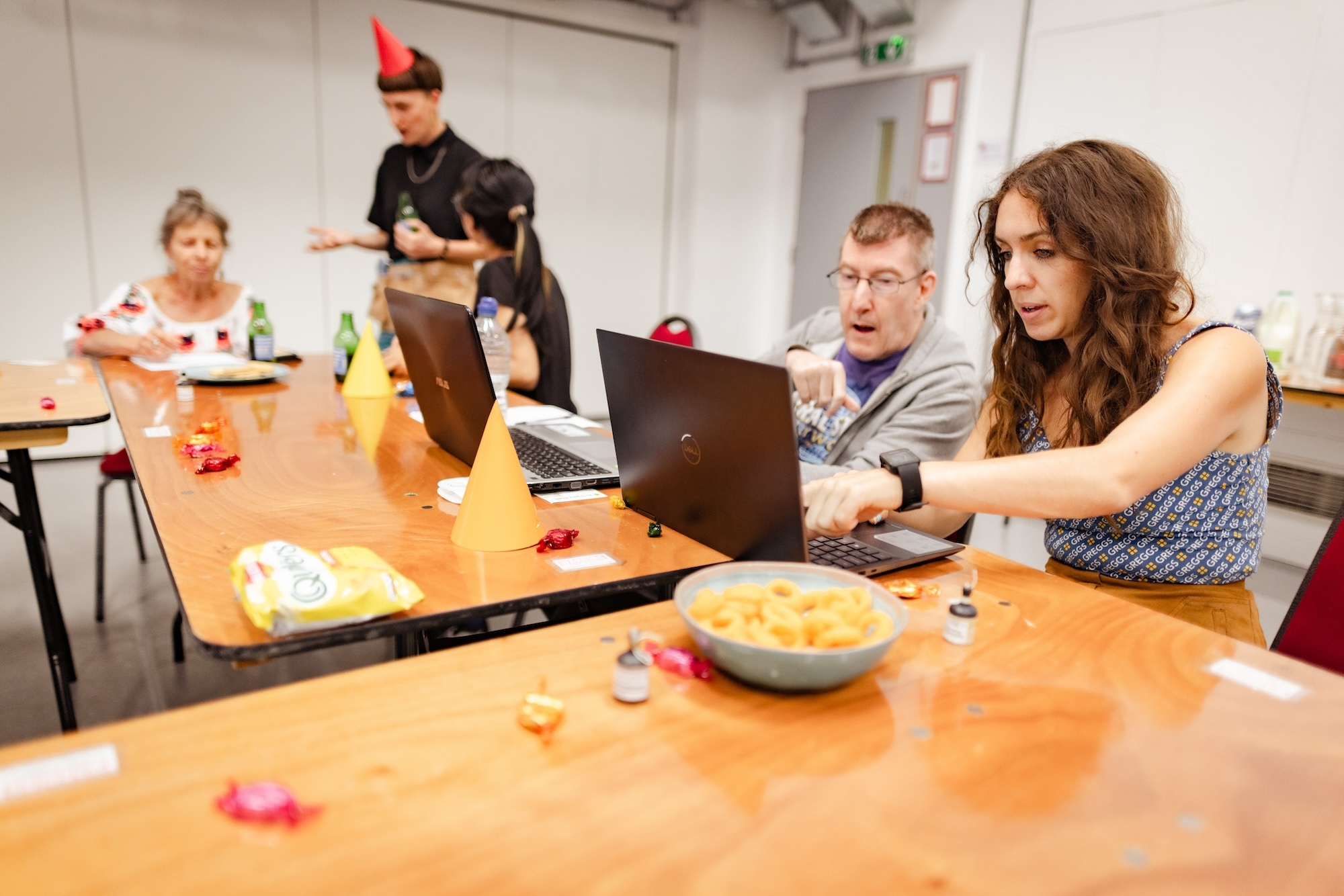 A group of people sits at a table with laptops, snacks, and drinks. Two in the foreground focus on their laptops while others chat in the background.