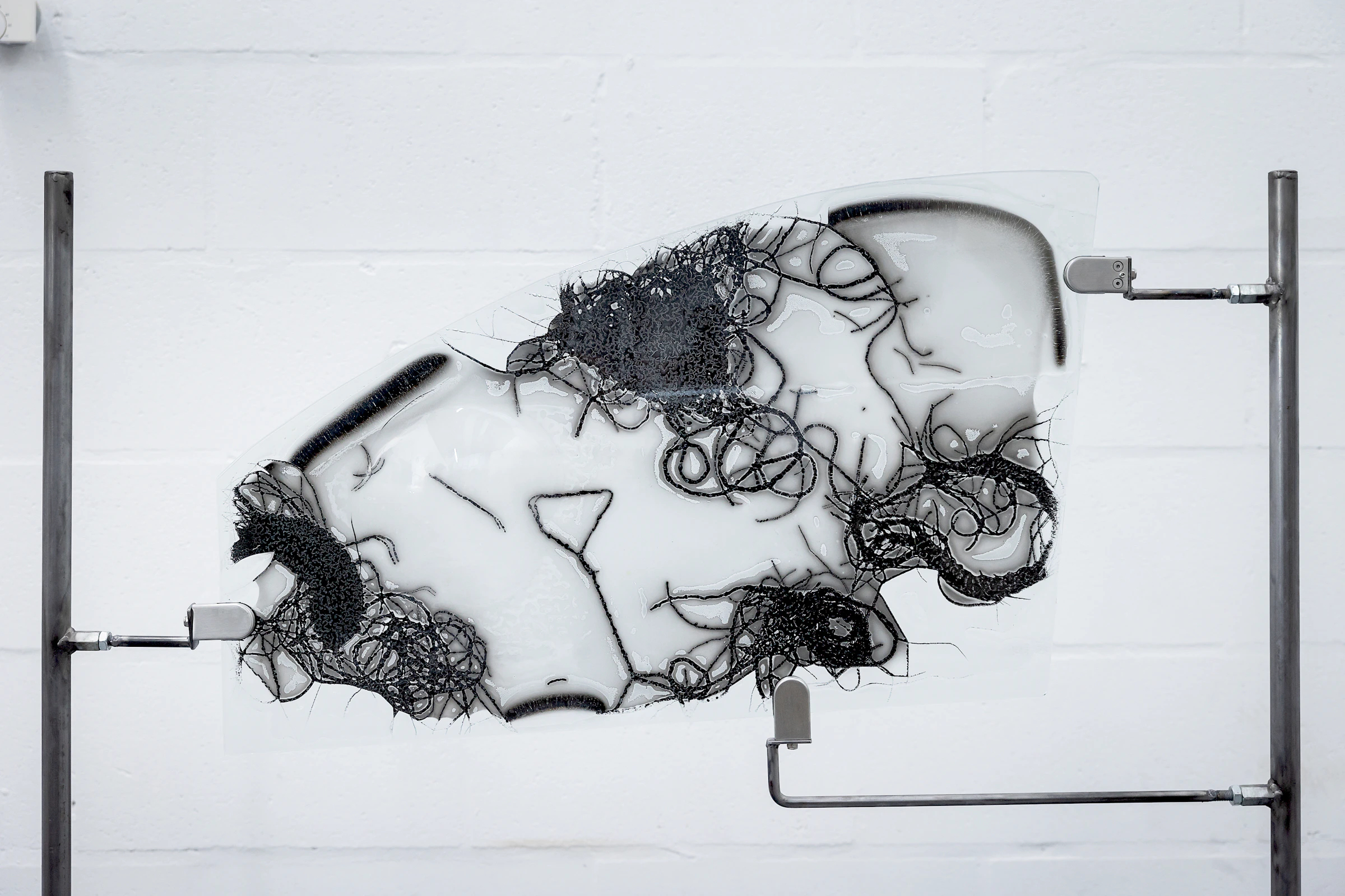 A sculpture presented against a white wall consisting of a glass panel in the shape of a car window with embedded horse hair and ash, which is held by two metal rods.