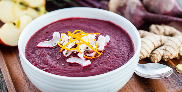 Beet soup with coconut flakes and orange zest served in a white bowl on a wooden tray. Ginger, beets and sliced apples in background.