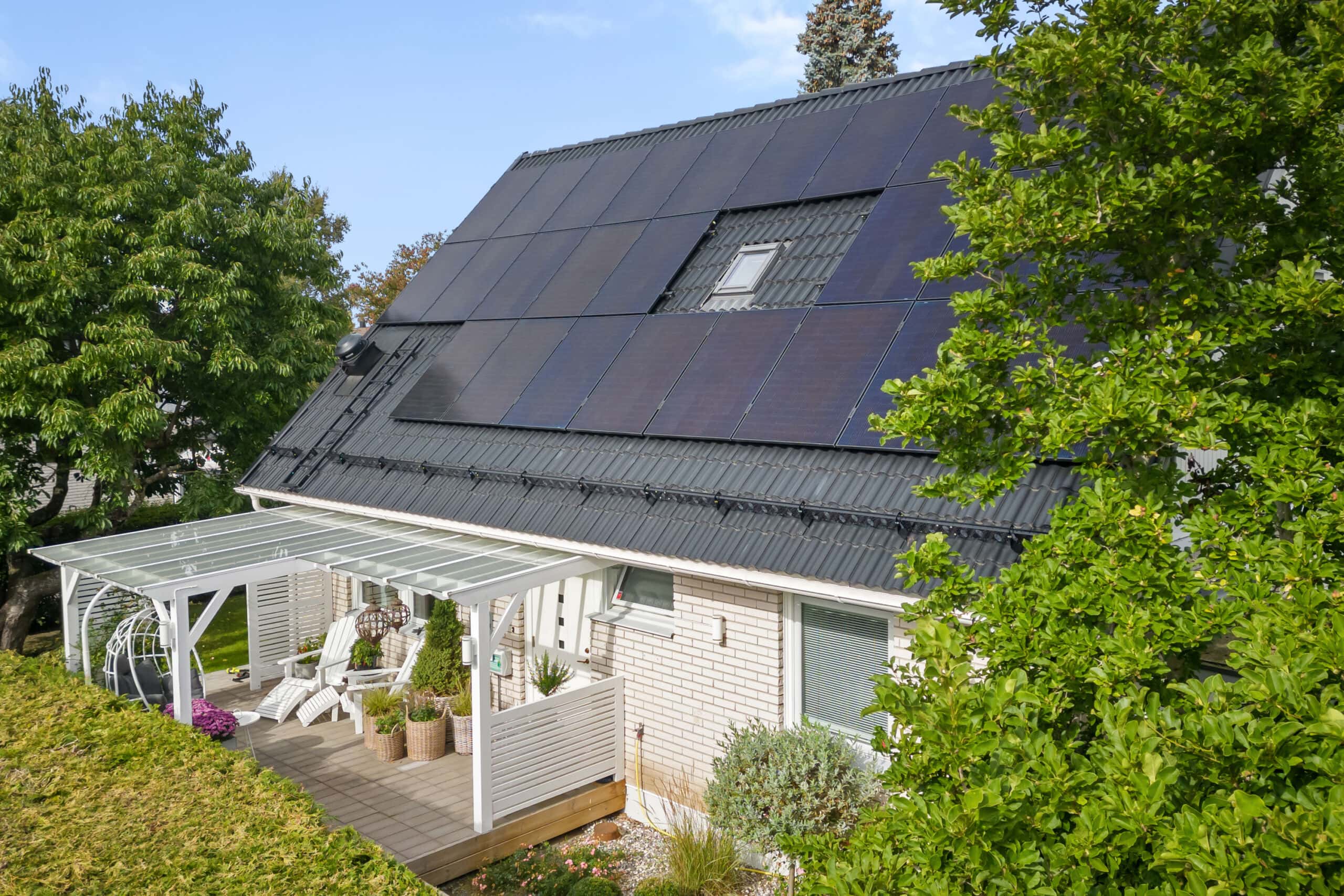 Svarta solceller på villa, klarbli himmel och fin pergola feamförhuset. Grön häck i förgrunden.